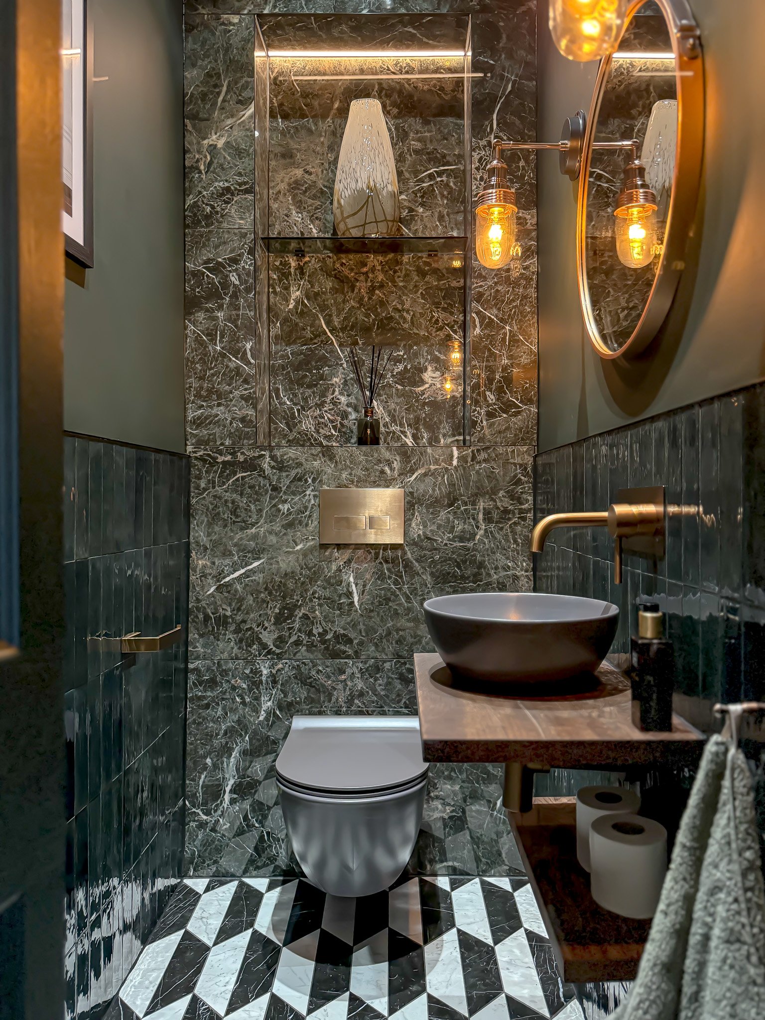 Luxury cloakroom bathroom in North London featuring dark marble-effect wall tiles, geometric patterned floor tiles, brass wall-mounted tapware, a stone countertop basin and warm statement lighting by North Arch Bathrooms.