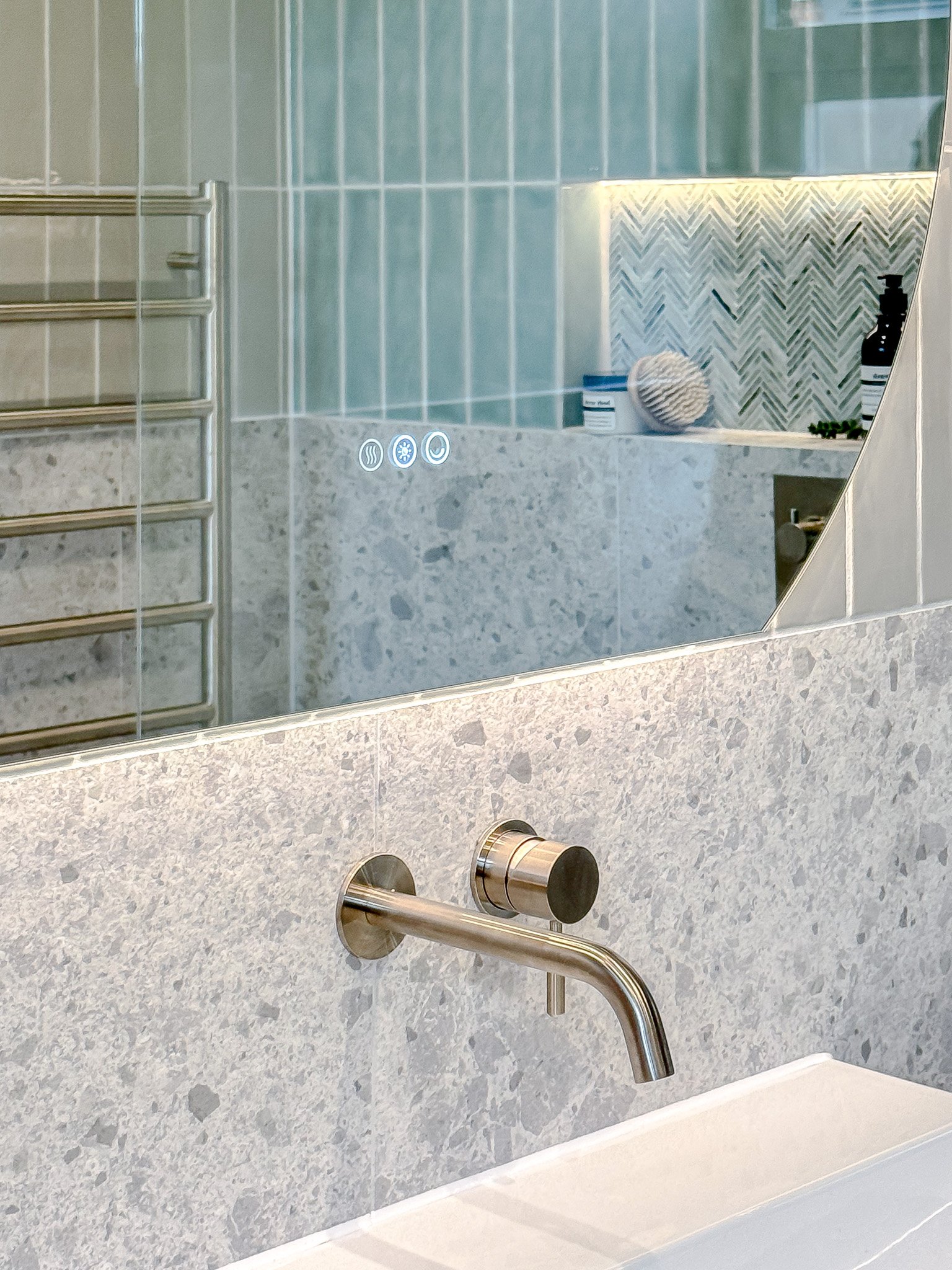 Bathroom mirror and vanity detail with terrazzo wall tiles, brushed metal wall-mounted tap and illuminated touch-control mirror