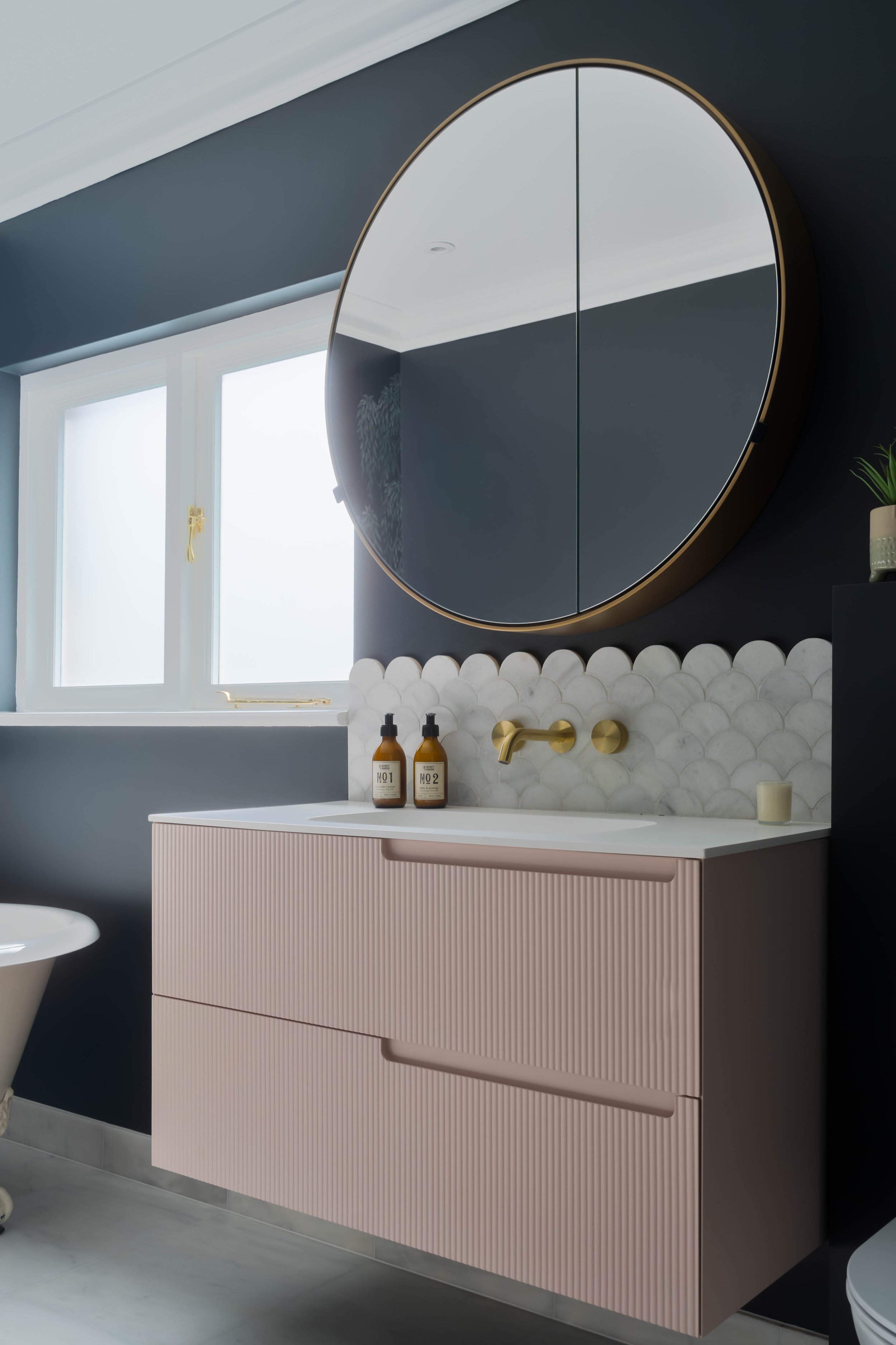Modern bathroom vanity with fluted blush cabinet, white countertop and brushed brass wall-mounted tap, set against white marble scallop tiles and a round mirrored cabinet with gold trim