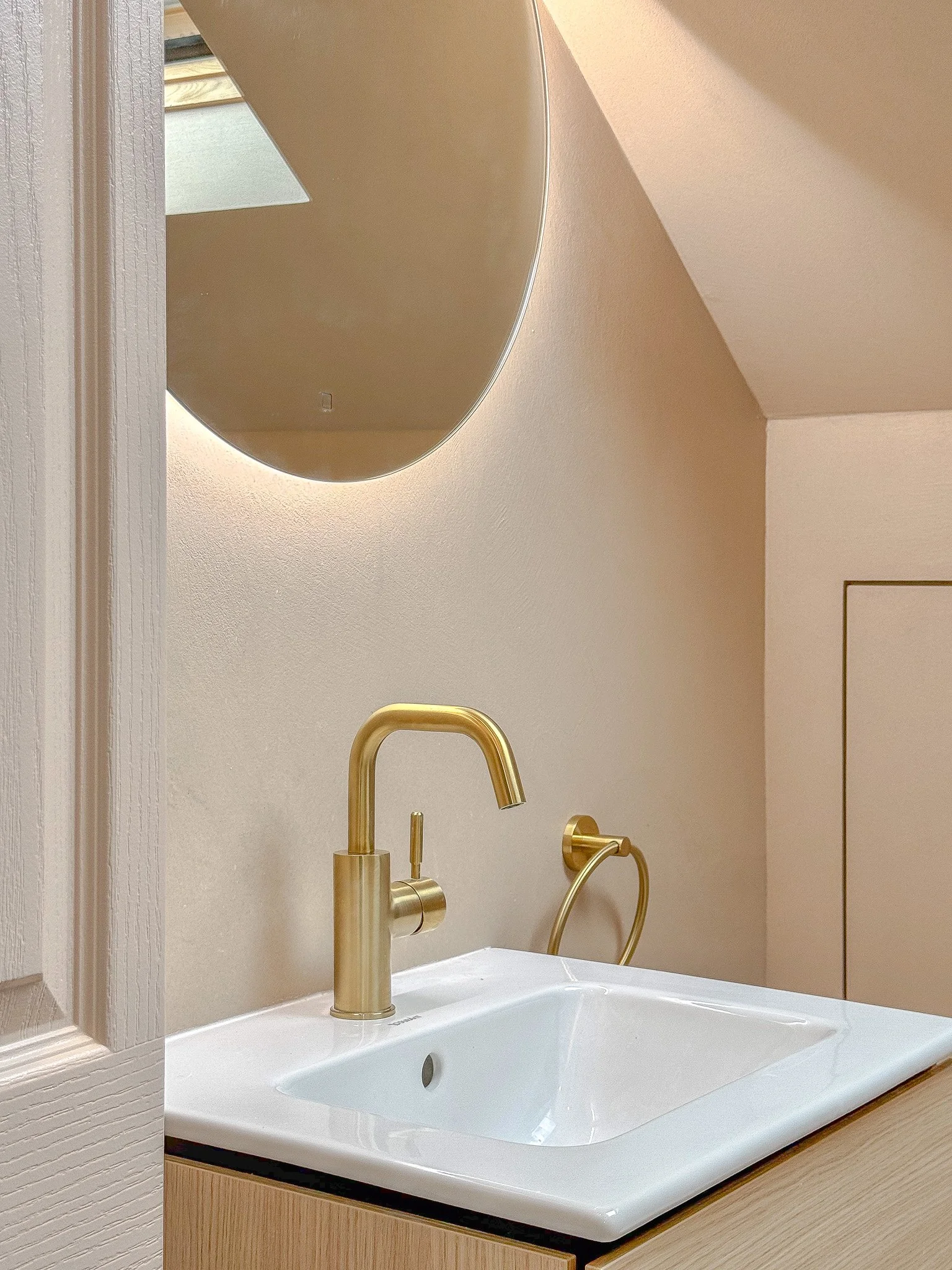 Luxury modern bathroom vanity with brushed brass basin mixer tap, integrated white ceramic sink, wall-mounted brass towel ring and backlit curved mirror, showcasing minimalist design and high-end bathroom finishes.