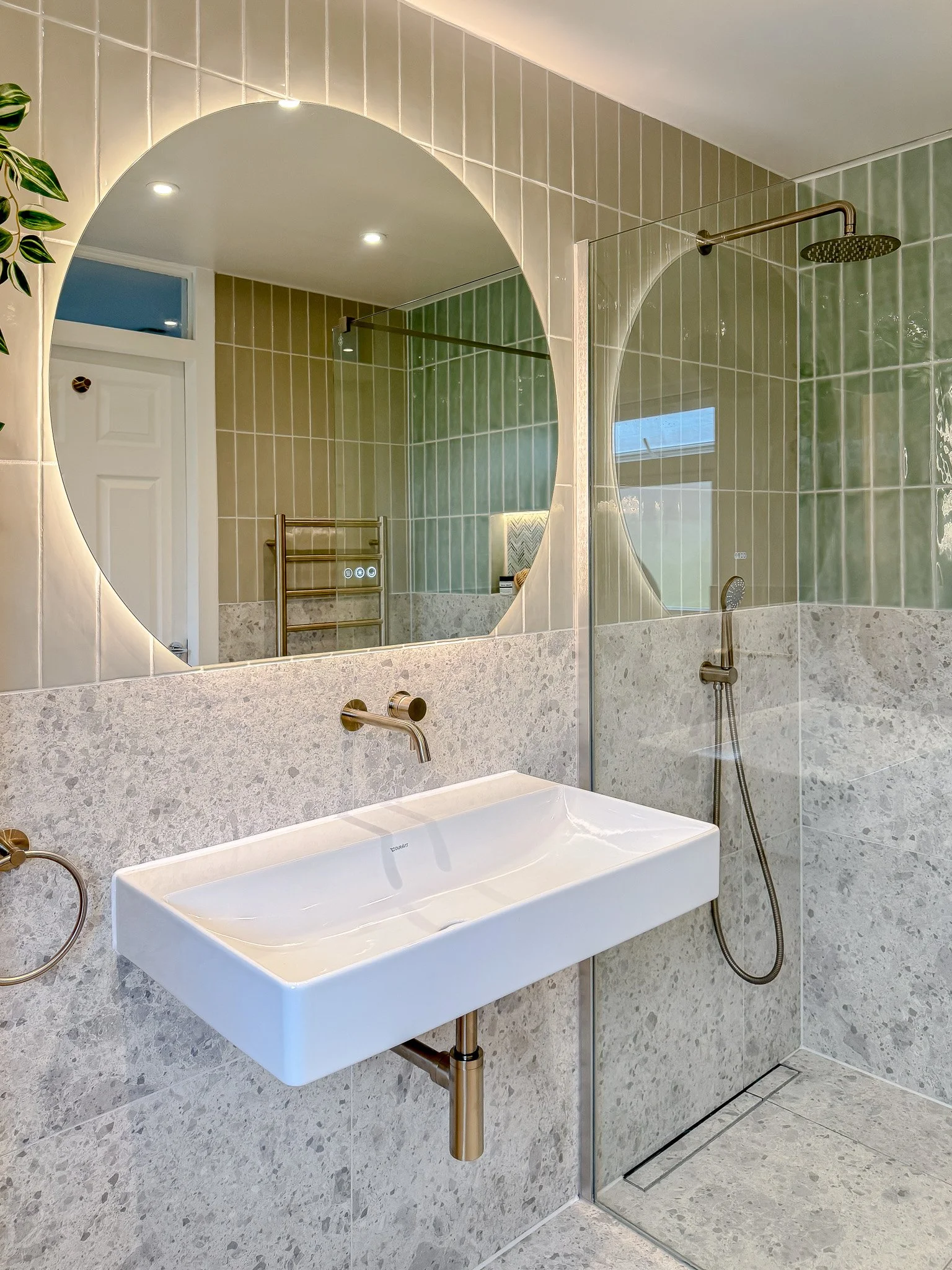 Modern bathroom with terrazzo stone wall tiles, vertical green ceramic tiles, a large round mirror and a wall-mounted brushed brass tap over a wide rectangular white basin.