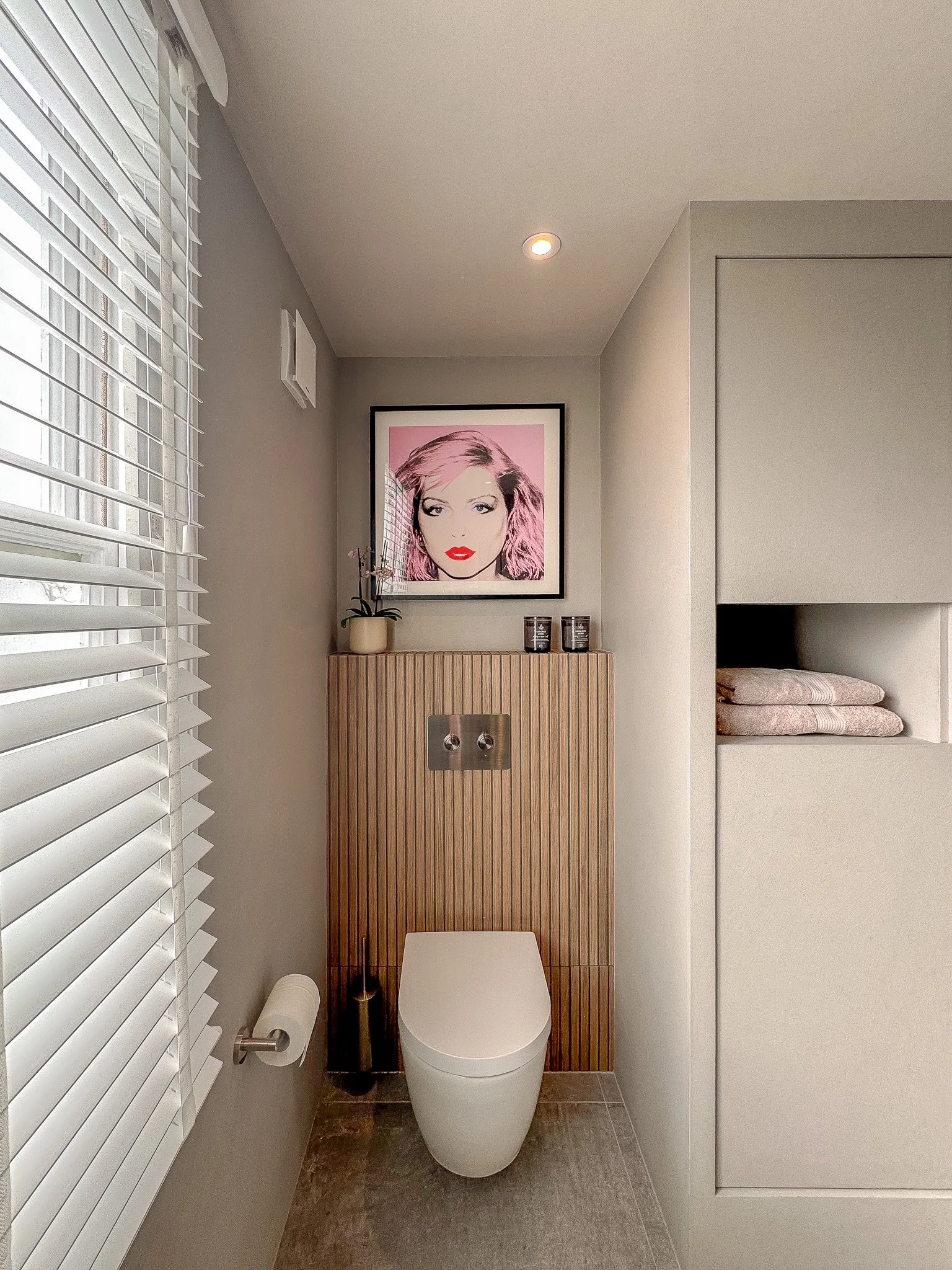 Contemporary bathroom with vertical fluted wood cladding behind a wall-mounted toilet, neutral built-in shelving and a framed artwork display.