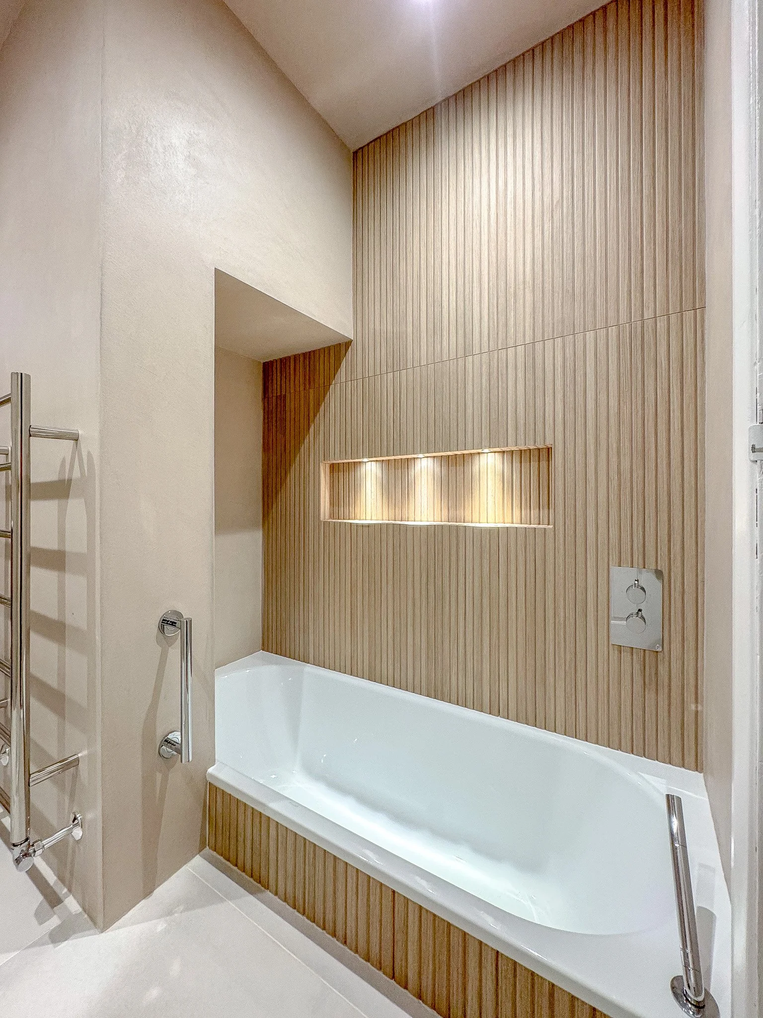 Contemporary bathroom in North London featuring a built-in bath with vertical wood-effect wall panelling, illuminated shower niche, minimalist chrome fittings and clean, modern finishes by North Arch Bathrooms.