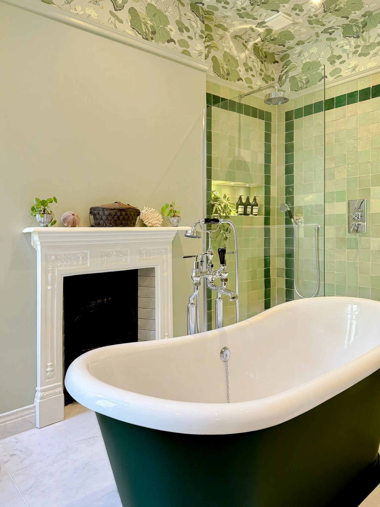 Luxury bathroom with a freestanding bathtub, green zellige tiles in a walk-in shower, decorative fireplace, and botanical wallpaper.