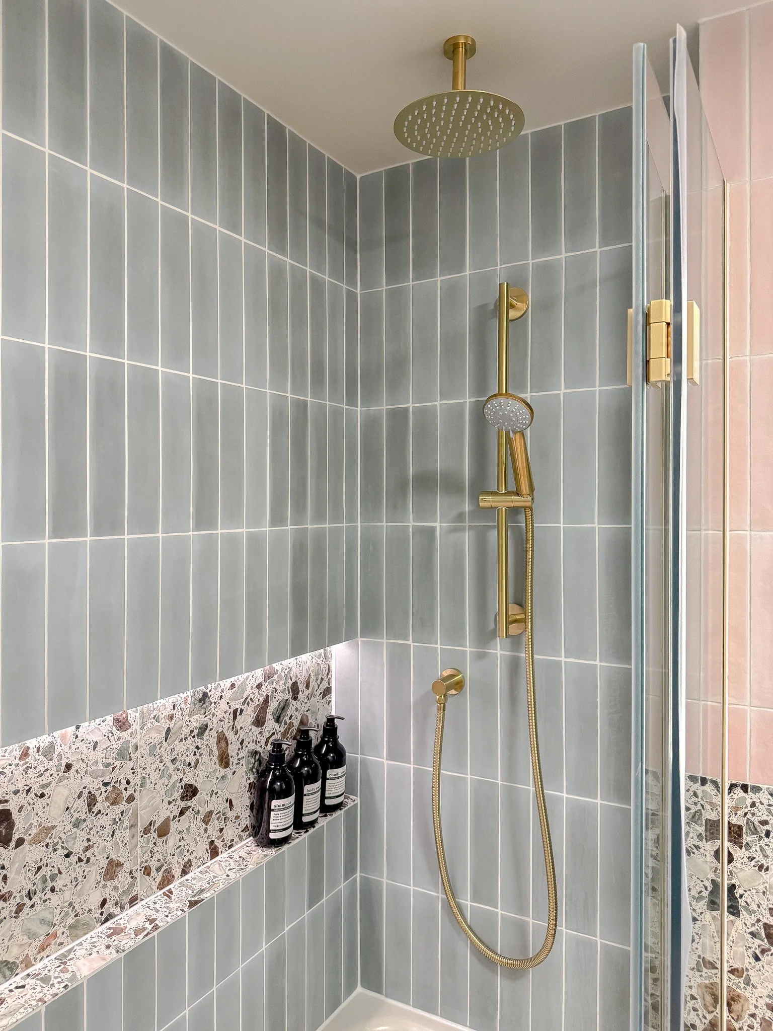 Contemporary walk-in shower with soft grey vertical tiles, brushed brass rainfall shower and terrazzo recessed niche in a bespoke luxury bathroom design