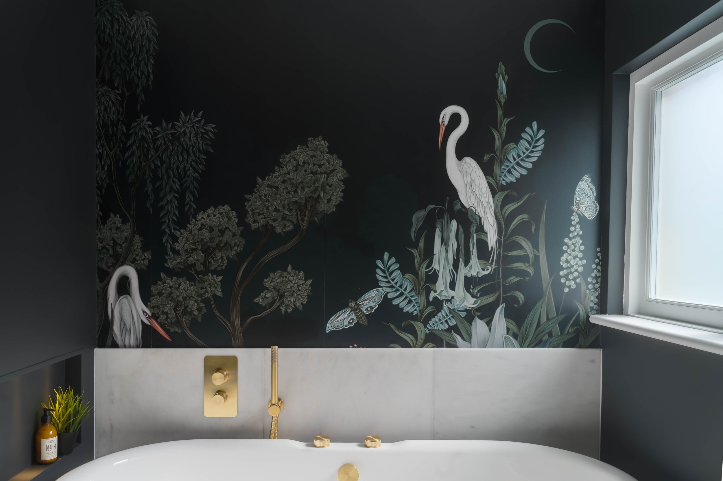 Luxury bathroom with marble splashback, brushed brass bath controls and handheld shower, featuring a dark botanical mural with heron and floral illustrations behind a freestanding tub