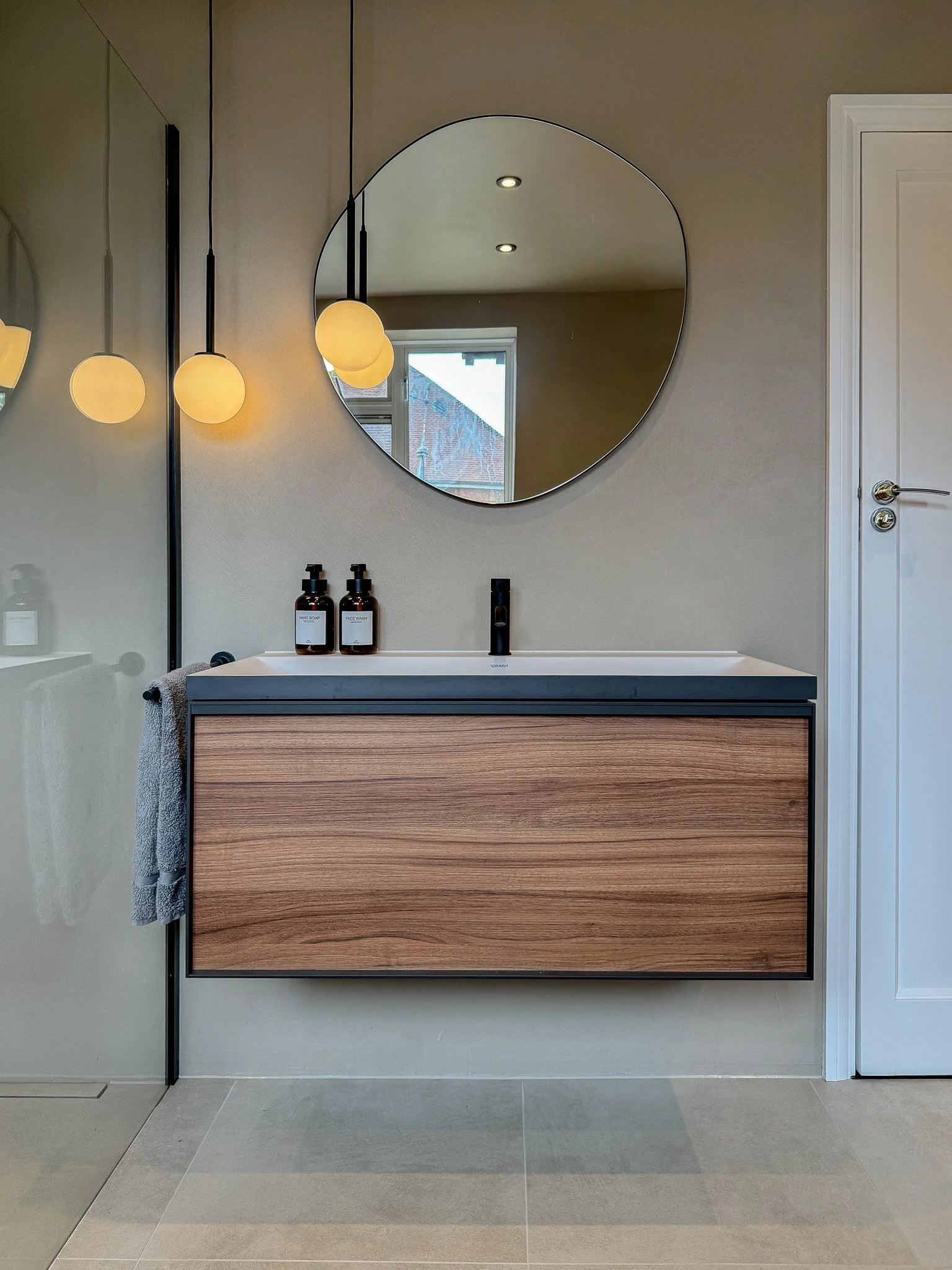 Contemporary bathroom design featuring a floating wood vanity, minimalist round mirror, soft globe pendants and matte black tapware.