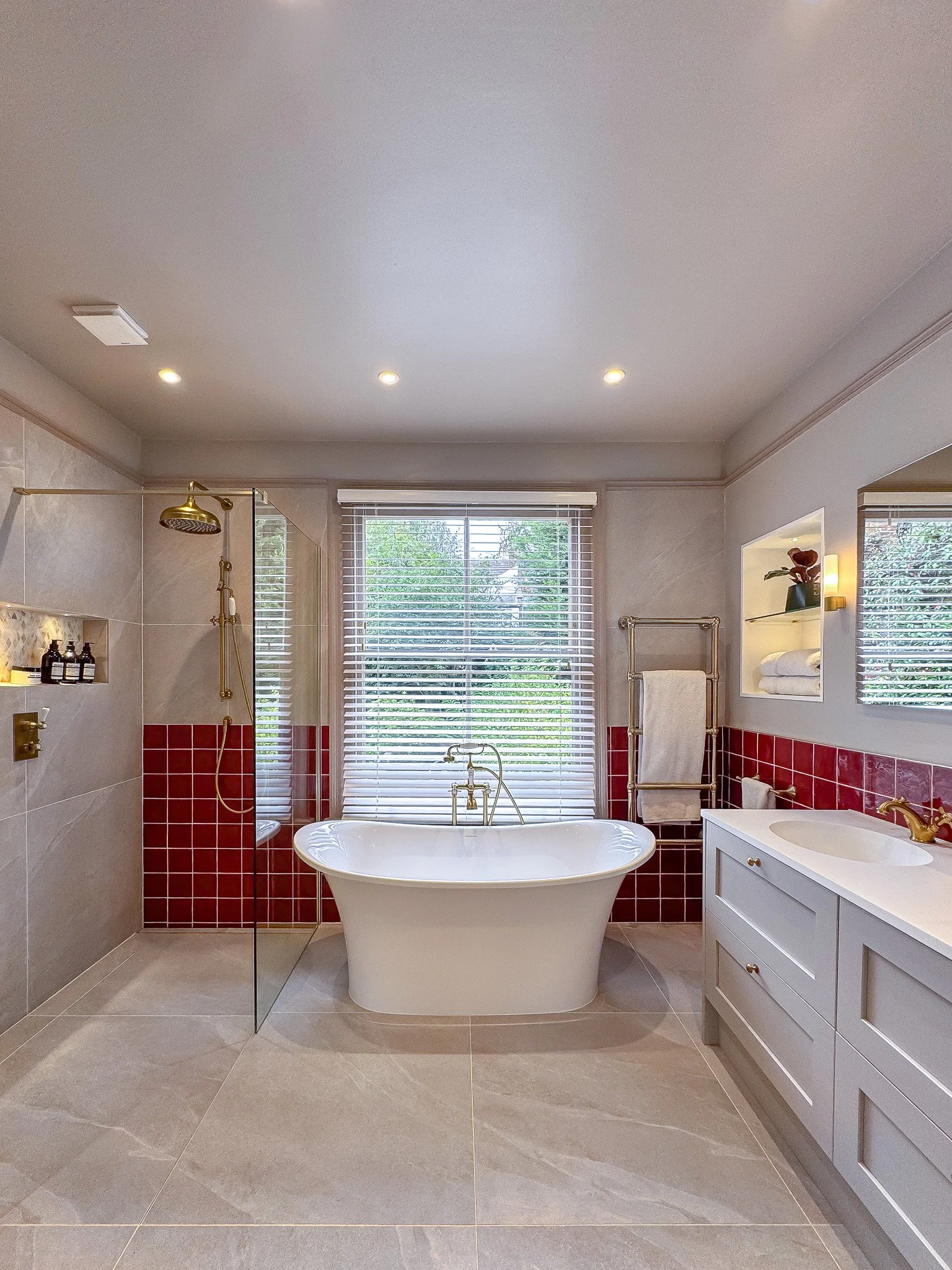 Bathroom with a freestanding bathtub, red square wall tiles, brass fixtures, and a white vanity unit beneath a window with blinds.