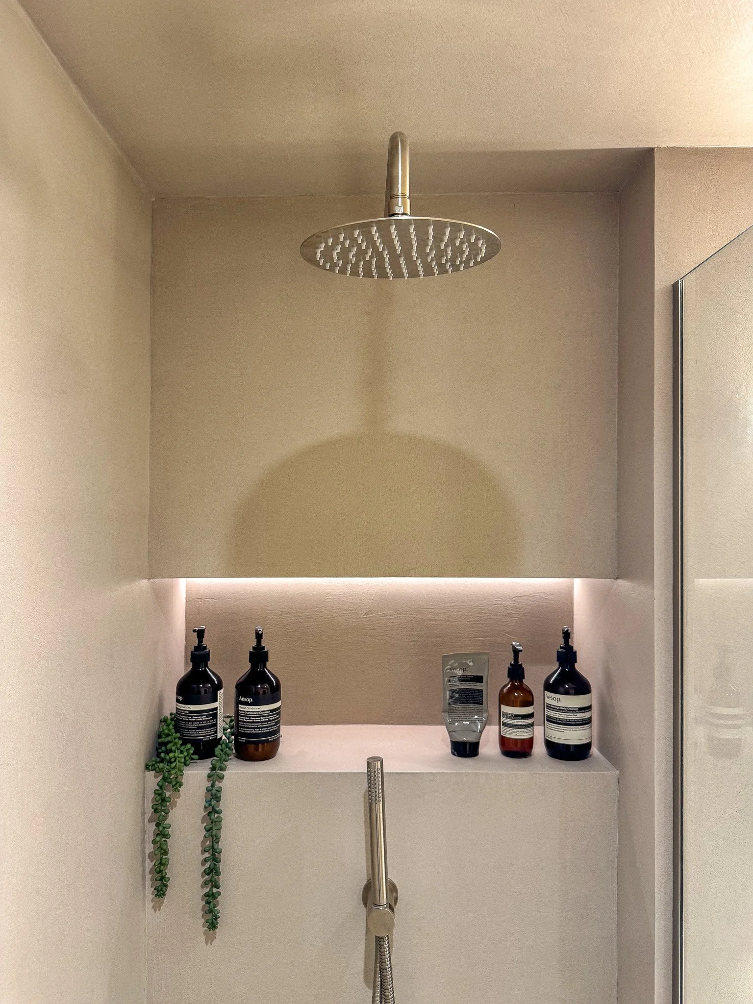 Minimalist walk-in shower with smooth microcement walls, stainless steel rainfall shower and recessed illuminated niche in a modern spa-style bathroom