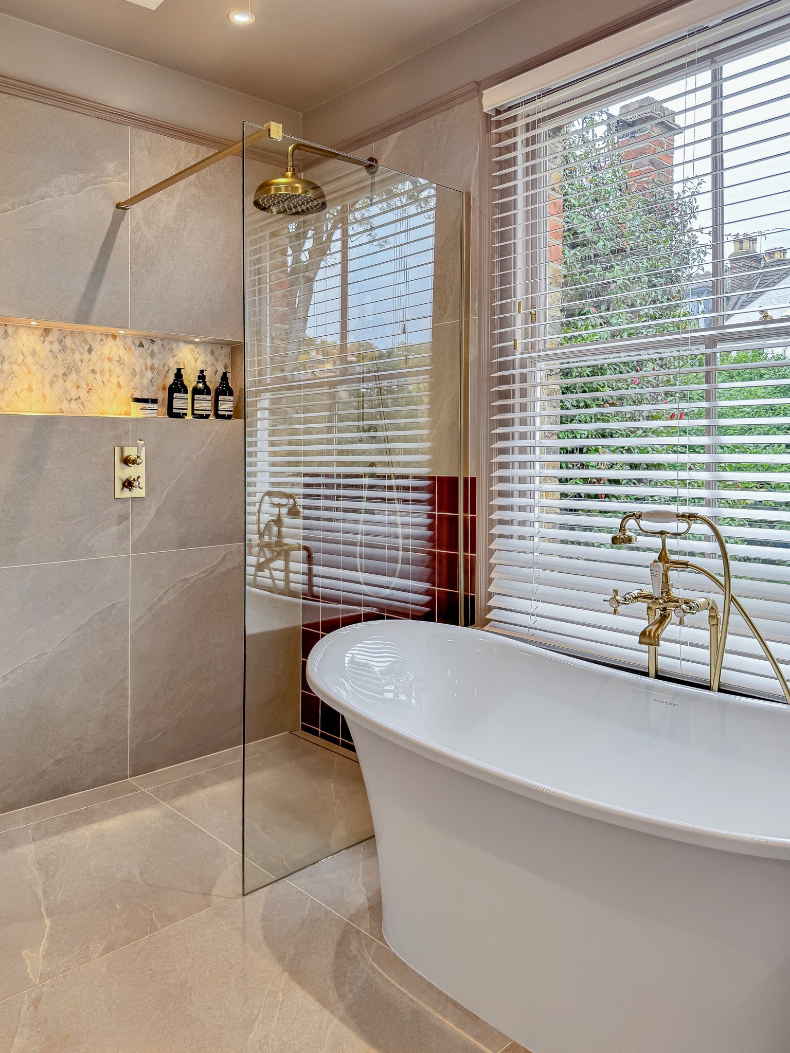 Modern bathroom featuring a freestanding bathtub with gold fixtures positioned near a large window with white blinds, and a separate shower area with beige tiles and a glass partition.