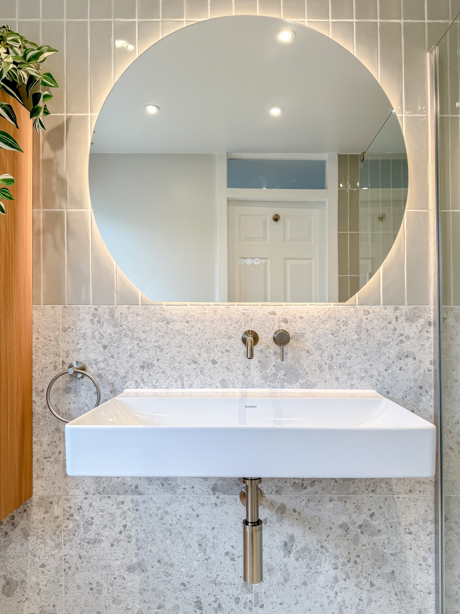 Contemporary bathroom vanity with wall-mounted basin, terrazzo splashback, brushed metal wall taps and large circular illuminated mirror.