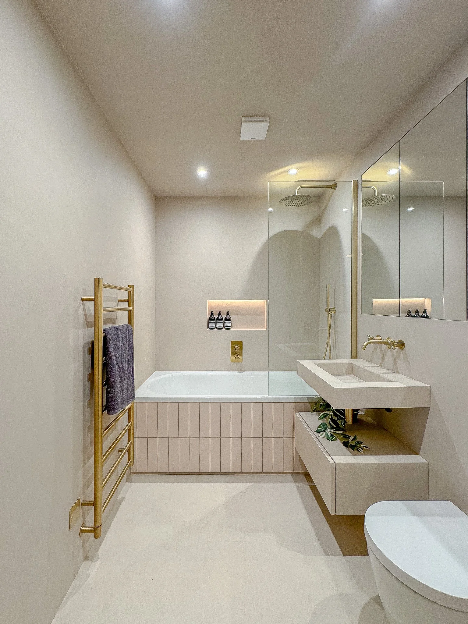 Modern luxury bathroom in North London featuring a built-in bath with tiled panel, brass fixtures, wall-mounted vanity, walk-in shower with rainfall head and neutral microcement-style finishes by North Arch Bathrooms.