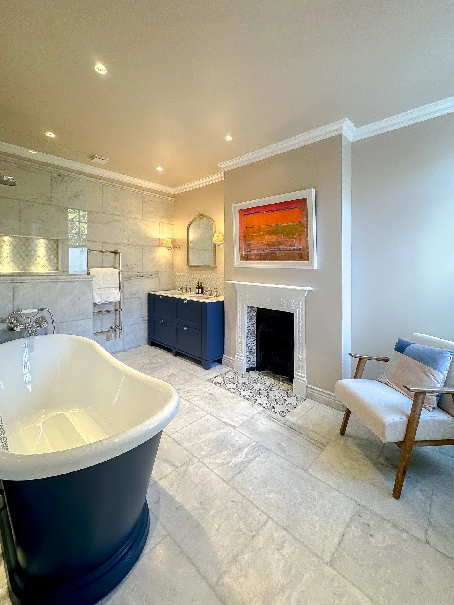 Classic luxury bathroom with freestanding bath, marble floor tiles, bespoke navy vanity unit and traditional fireplace in a design-led London home