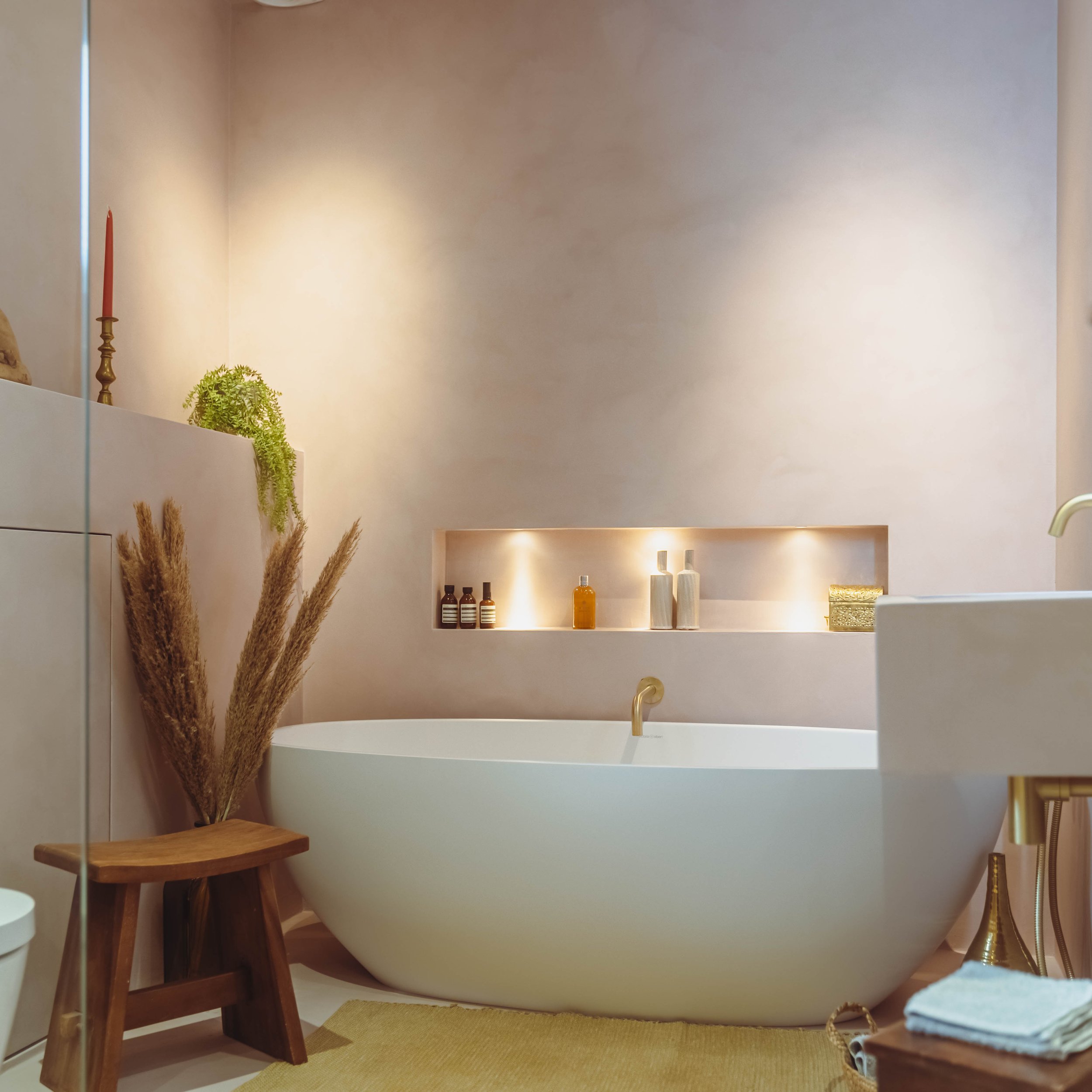 Calming spa-style bathroom with freestanding white bathtub, brushed brass tap, microcement walls and illuminated built-in niche shelving displaying toiletries. Neutral tones with natural décor including pampas grass and wooden stool