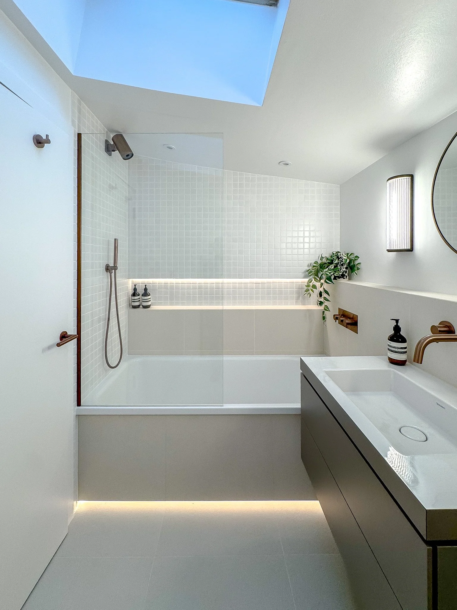 Minimalist bathroom with soft white tiles, integrated bathtub, brushed brass fixtures, illuminated niche shelving and a wall-mounted vanity sink.