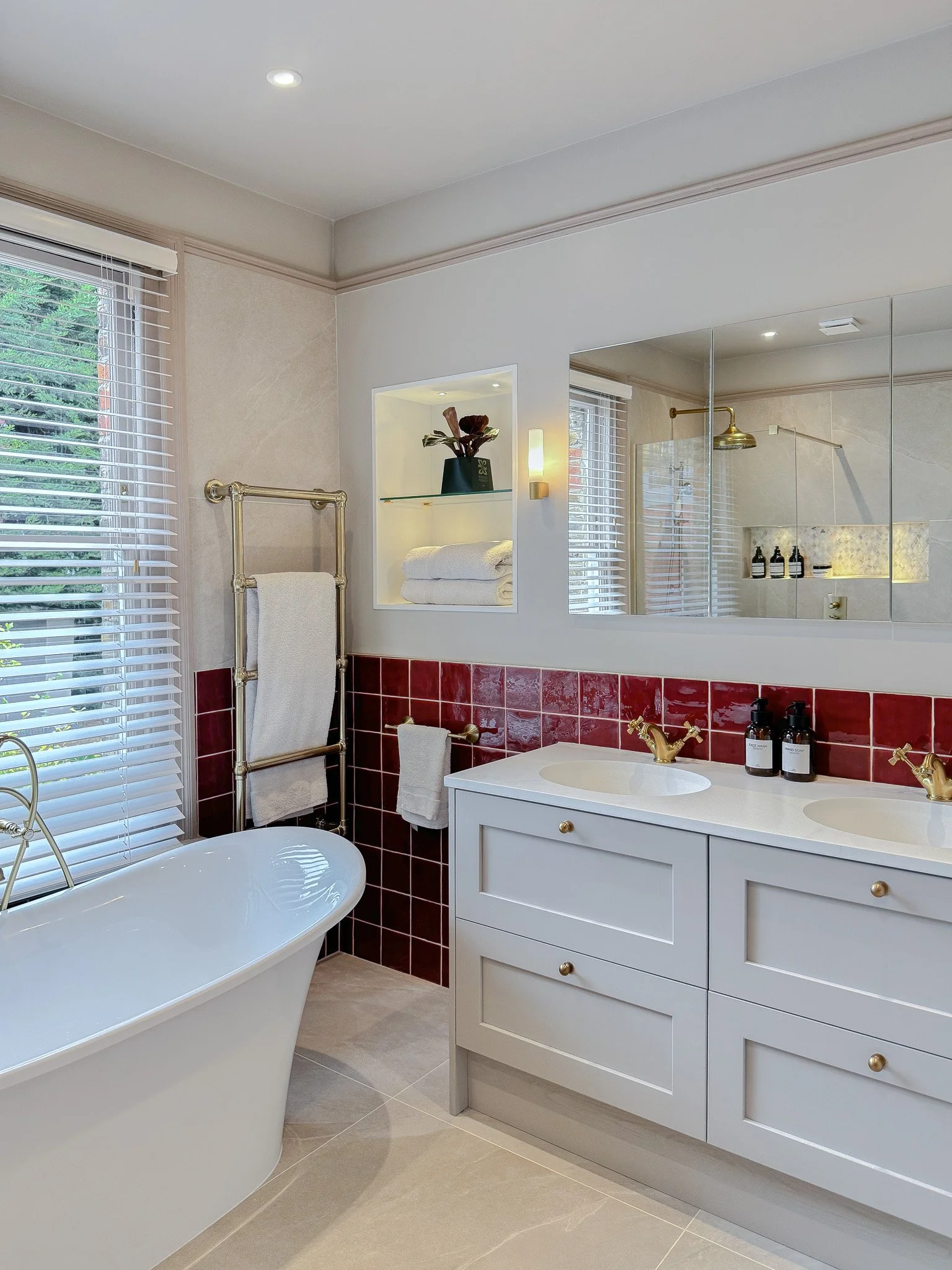 Modern bathroom with freestanding tub, brass fixtures, double vanity and rich red wall tiles, featuring built-in shelving and a large mirrored cabinet
