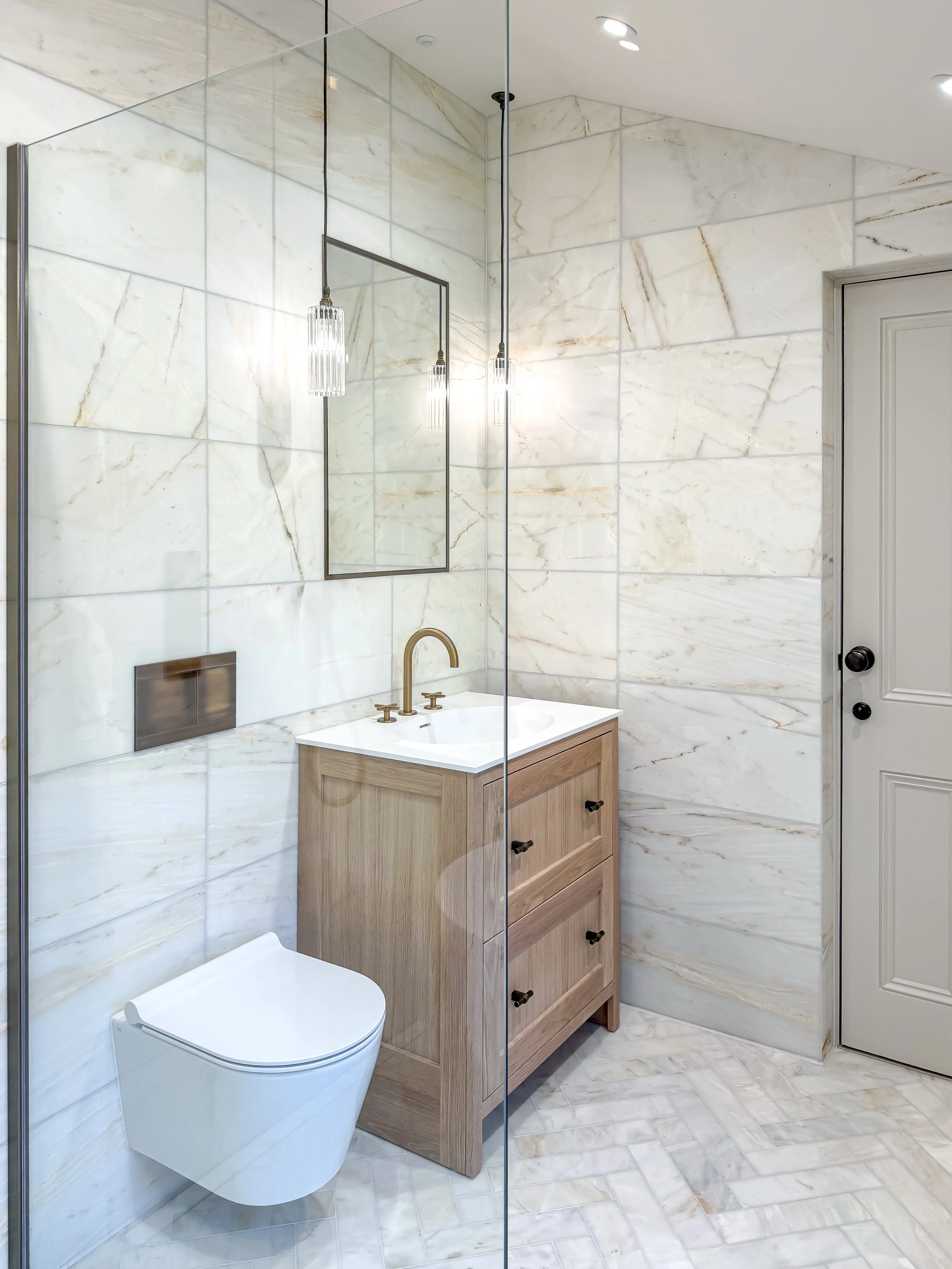 Luxury bathroom vanity in a North London home featuring marble-effect wall tiles, a timber vanity unit with white countertop basin, brushed brass tapware, framed mirror and statement pendant lighting by North Arch Bathrooms.