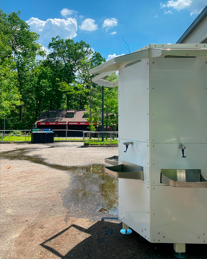 A woman filling a grey tumbler with water from an outdoor water refill station decorated with palm trees and a speaker illustration.