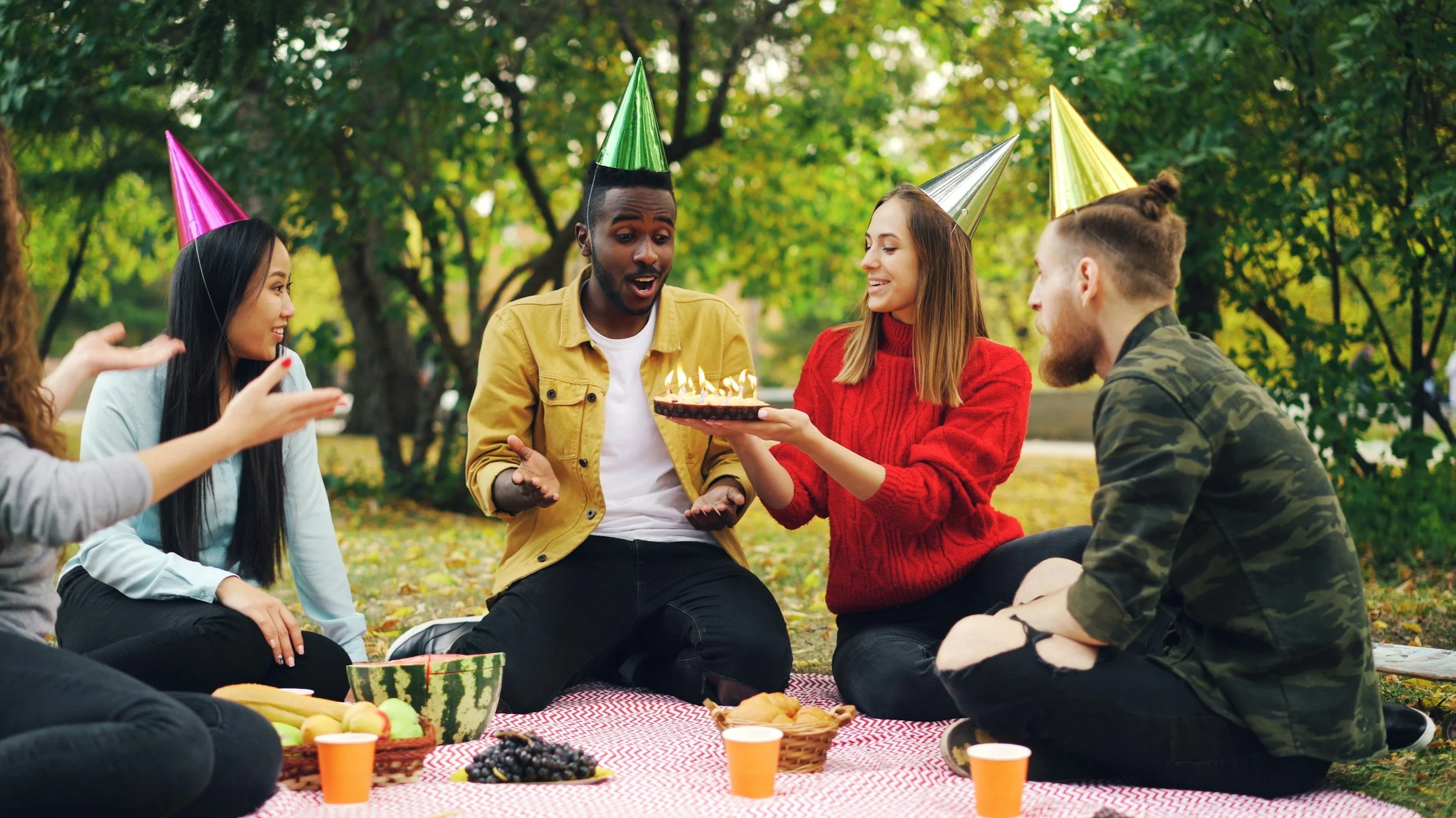 two people celebrating a birthday with hats, cake, candles, beverages, in a park setting