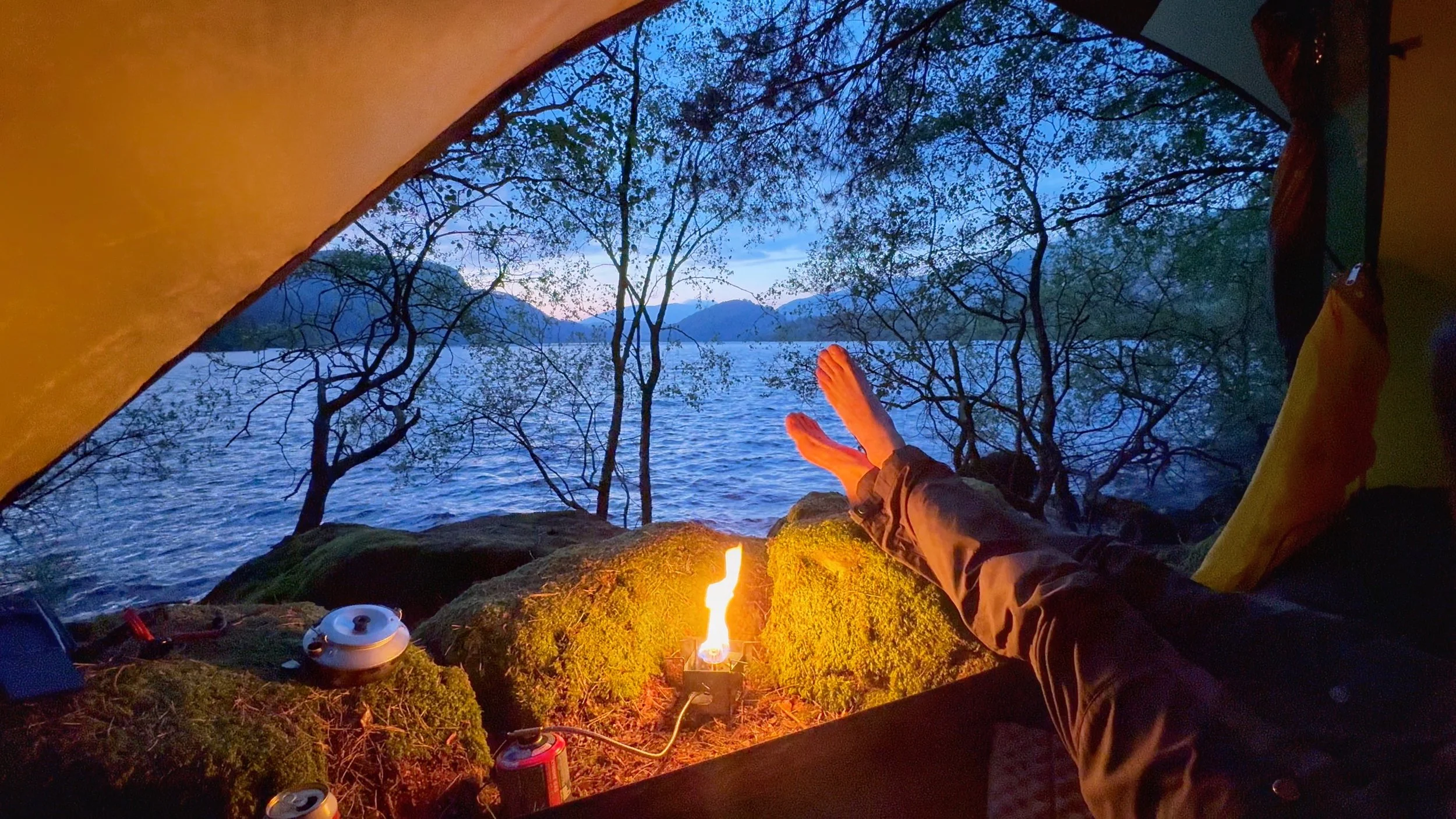 View from inside a tent looking out at a lake, trees, and mountains at sunset. A small fire is burning outside, with a person’s legs shown, and camping gear like a kettle nearby.