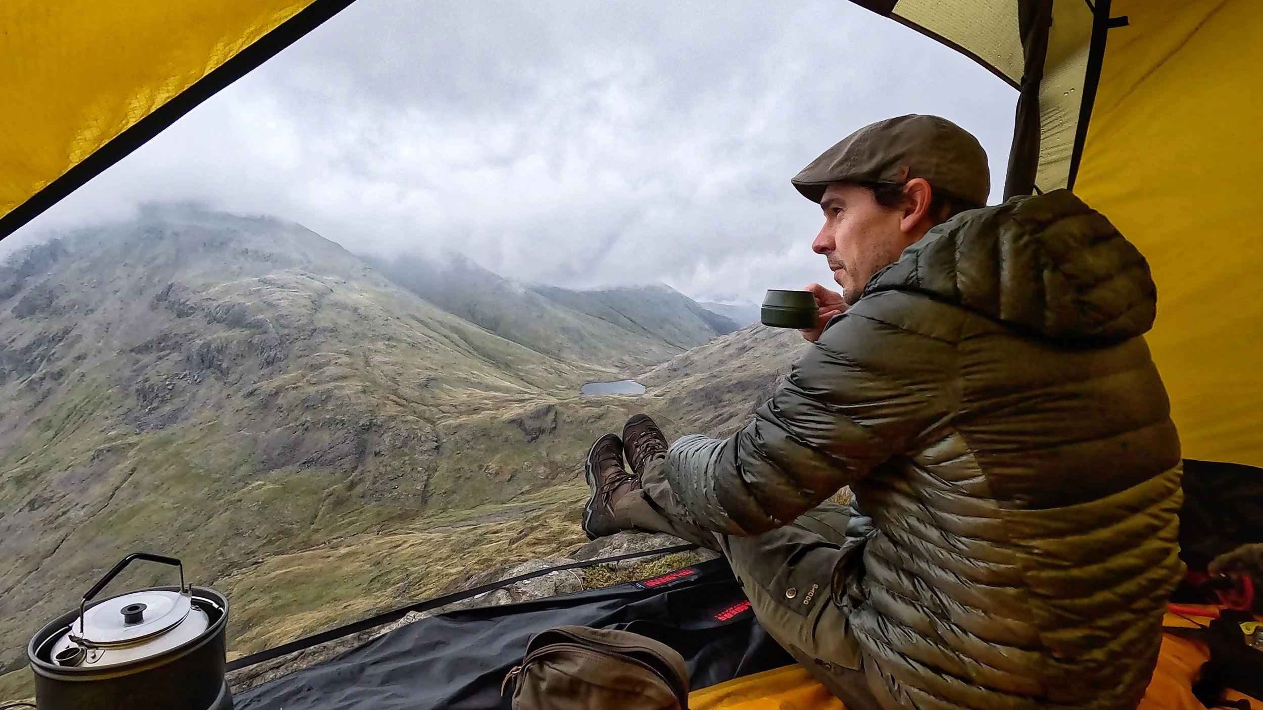 Person in a tent, sitting down with a mug, overlooking a misty mountainous landscape.