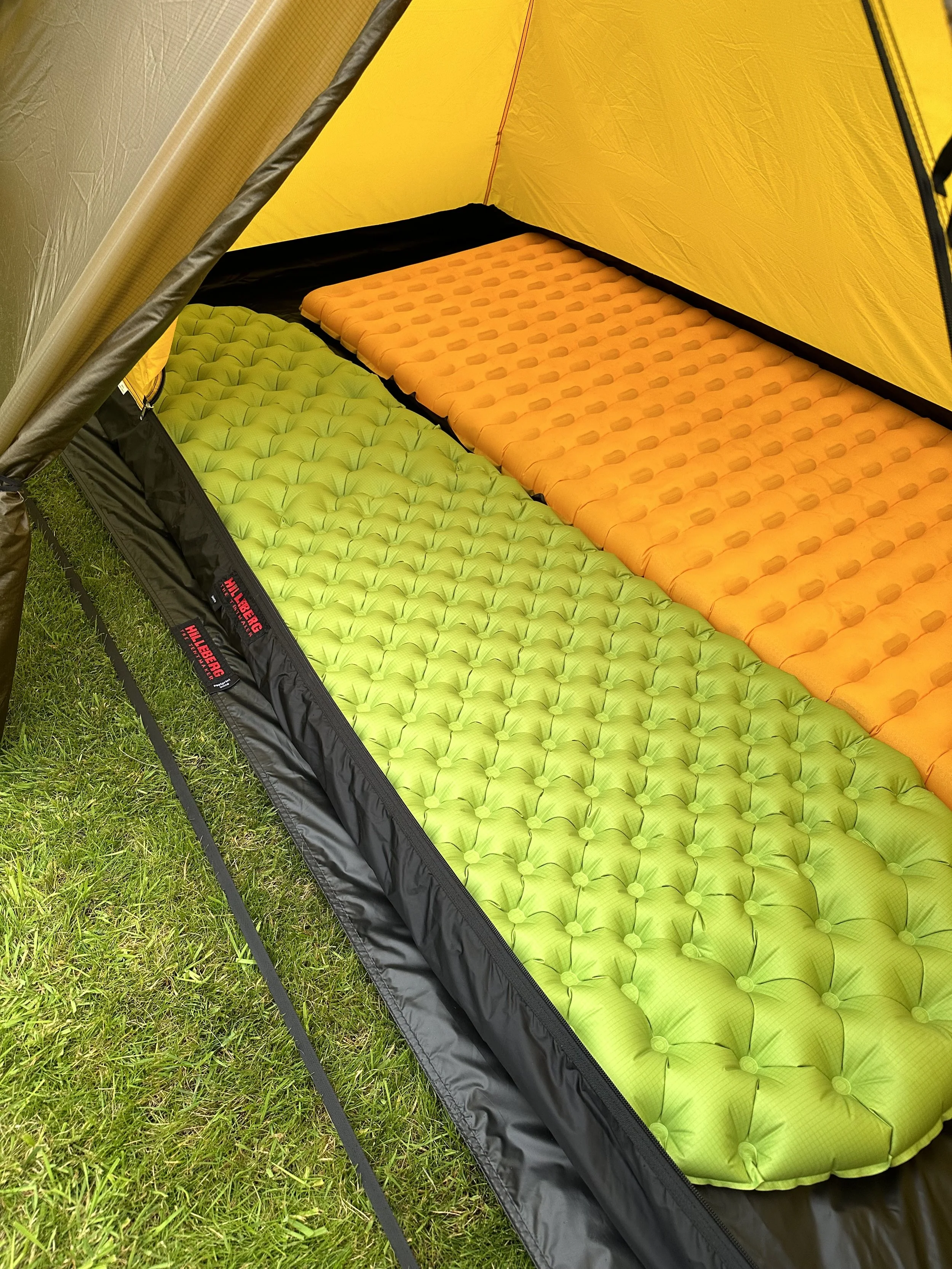 Two inflatable sleeping pads inside a yellow tent, one green and one orange, placed on the tent floor. The tent is set up on grass.