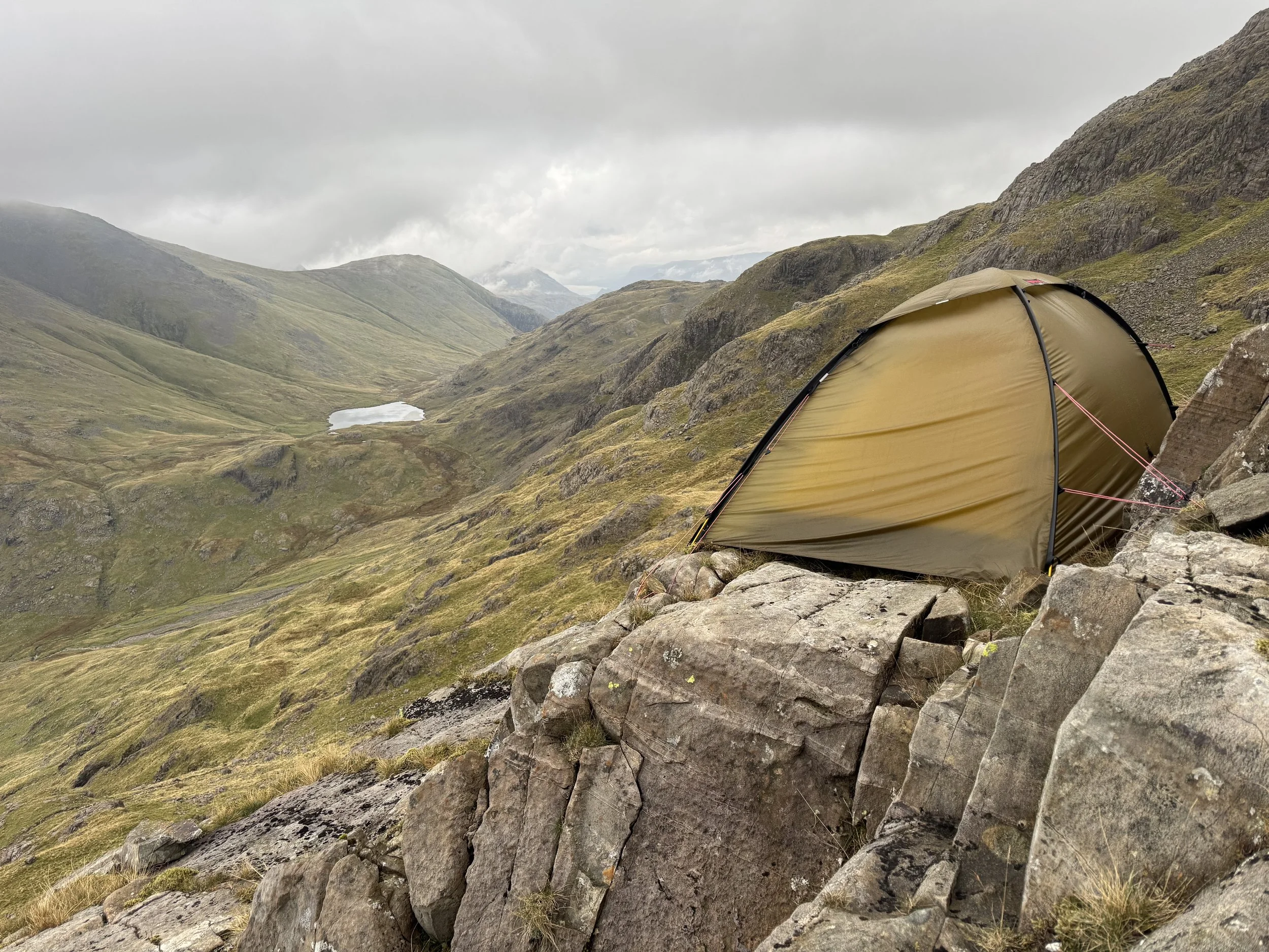 A camping tent set up on rocky terrain in a mountainous landscape, with grassy hills and a small lake in the distance under a cloudy sky.