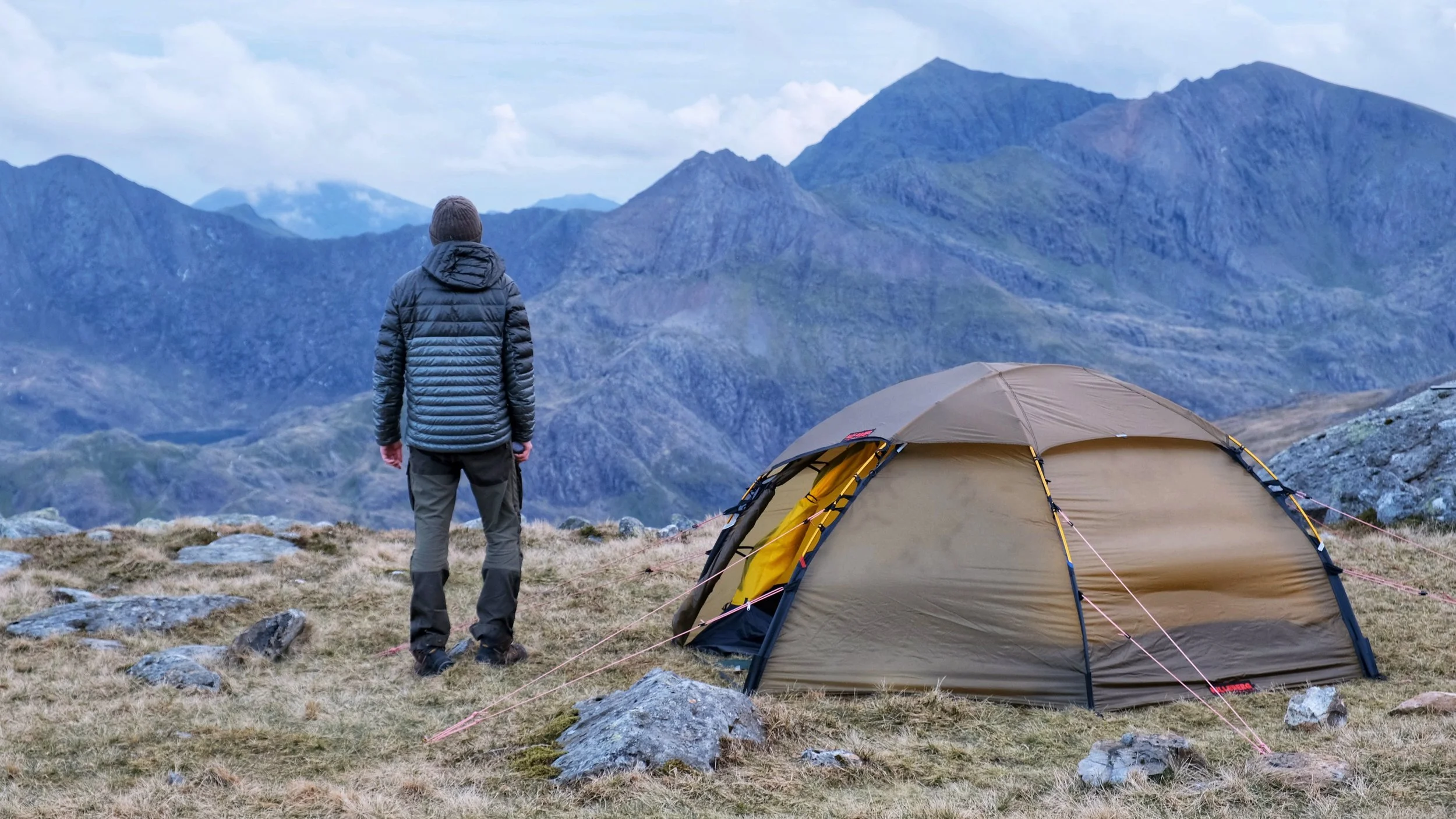 Person in a puffy jacket standing near a tent on a mountainside, overlooking a mountain range.