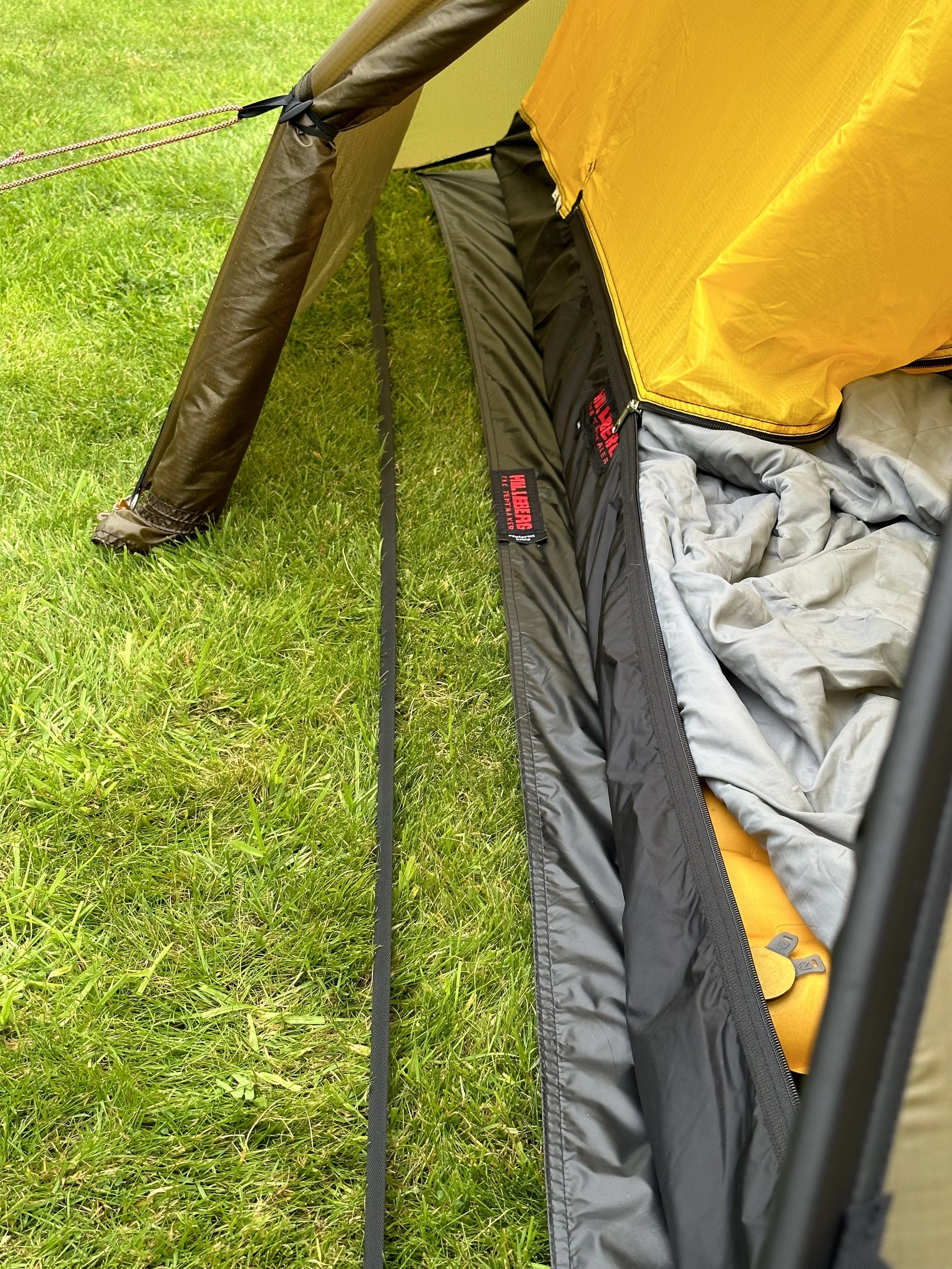 Close-up of a partially opened tent on grass, showing the tent's sidewall, groundsheet, sleeping bag or pad inside, and ropes securing the tent’s structure.