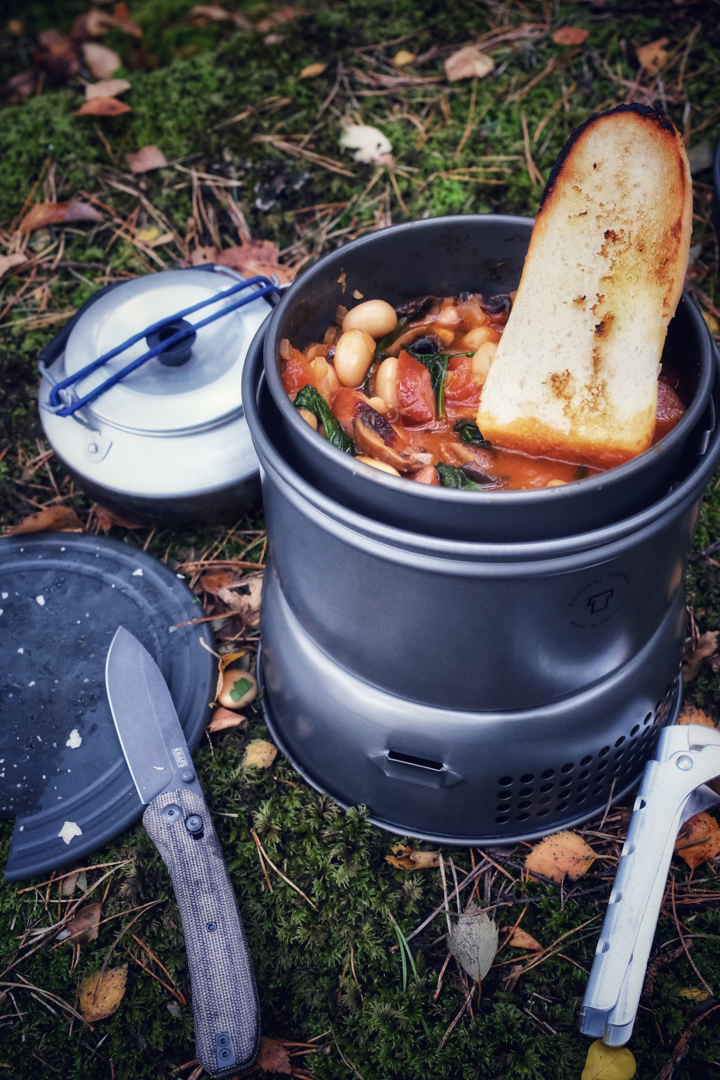 Outdoor cooking pot with sausages, mushrooms, beans, spinach, and tomato sauce, topped with grilled bread slice.
