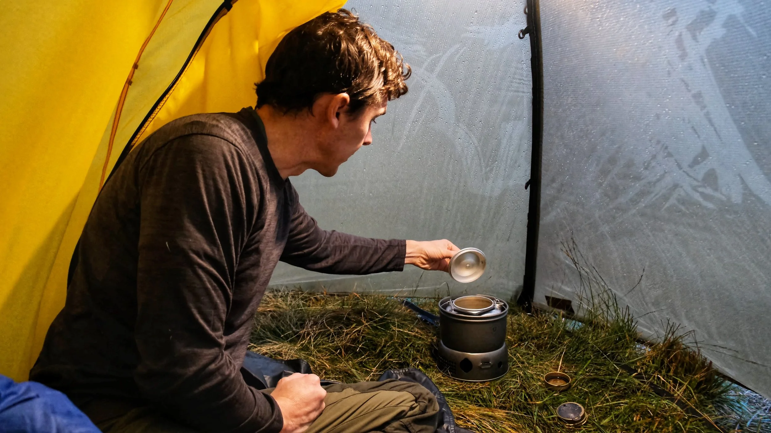 Person inside a yellow tent, using a portable camping stove on grass, holding a lid.