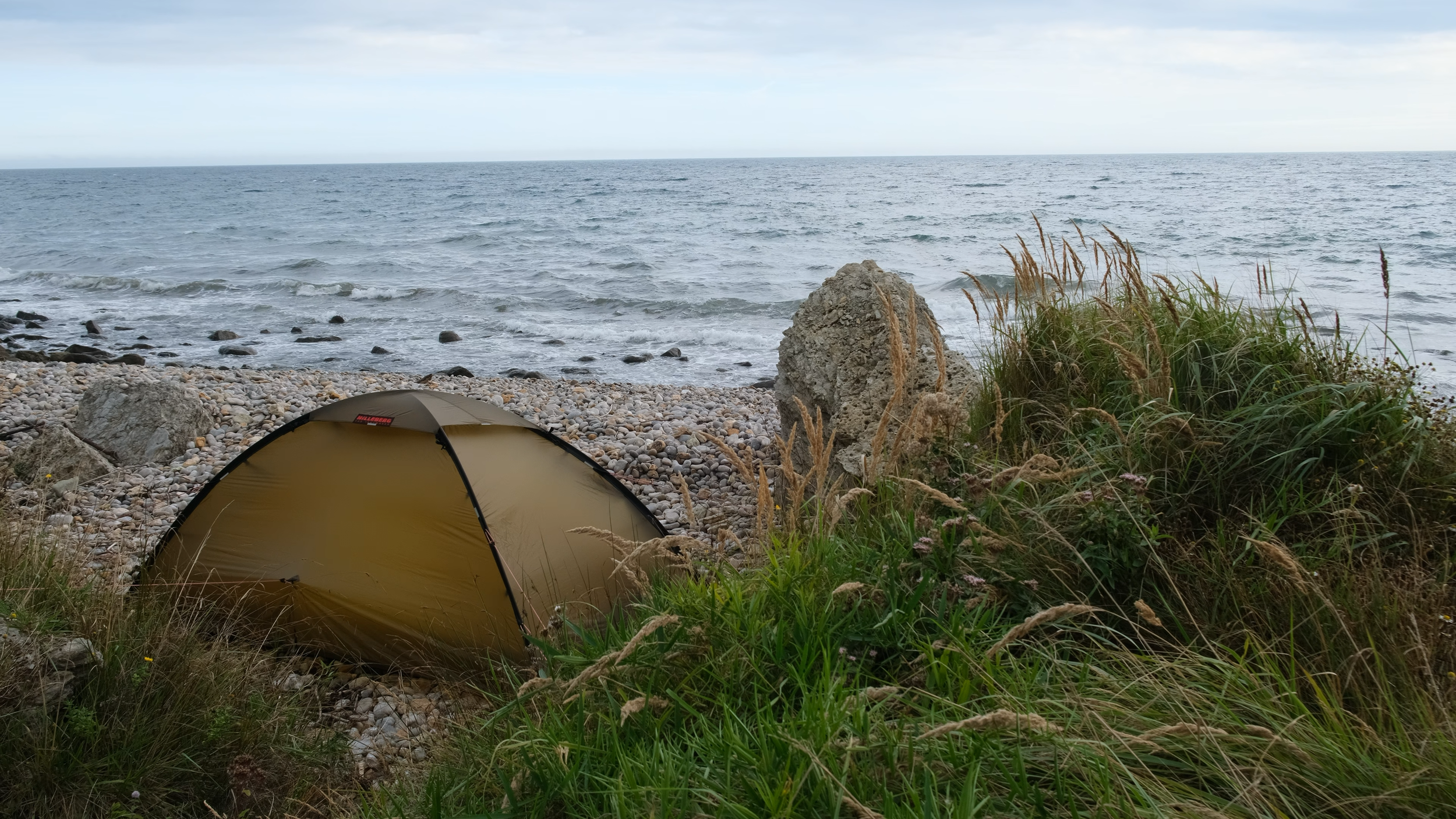 A brown tent pitched on a rocky beach with grassy areas, near an ocean with waves and cloudy sky.