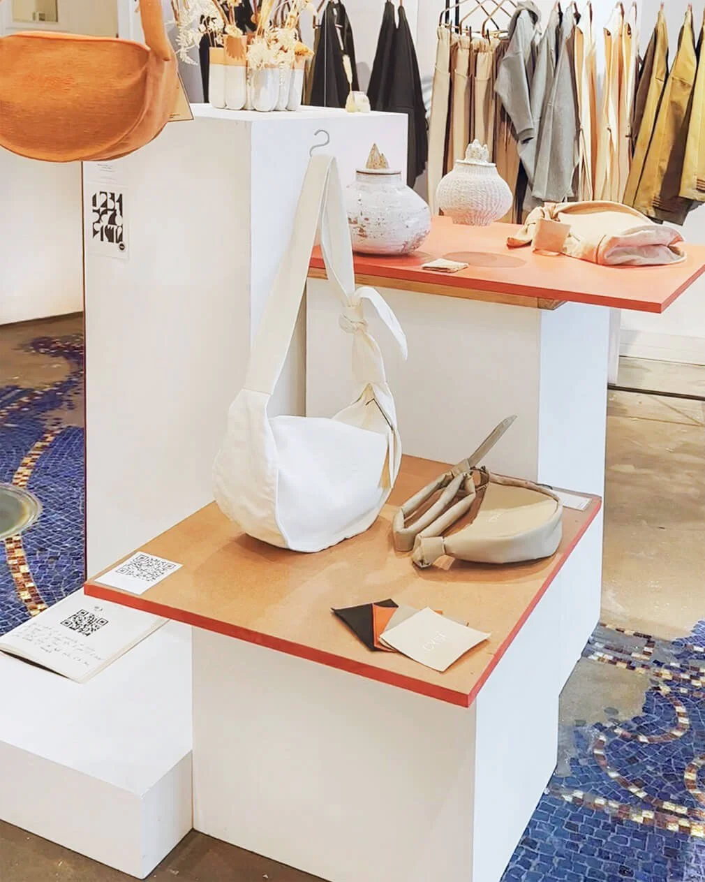 Affichage de vêtements et d'accessoires dans une boutique, avec des sacs, vases, et des vêtements suspendus, sur un fond de sol en mosaïque colorée.