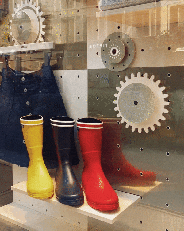 Des bottes de pluie jaunes, noires et rouges sont exposées devant un mur en miroir avec des engrenages en décor. Un sac bleu est suspendu à gauche.