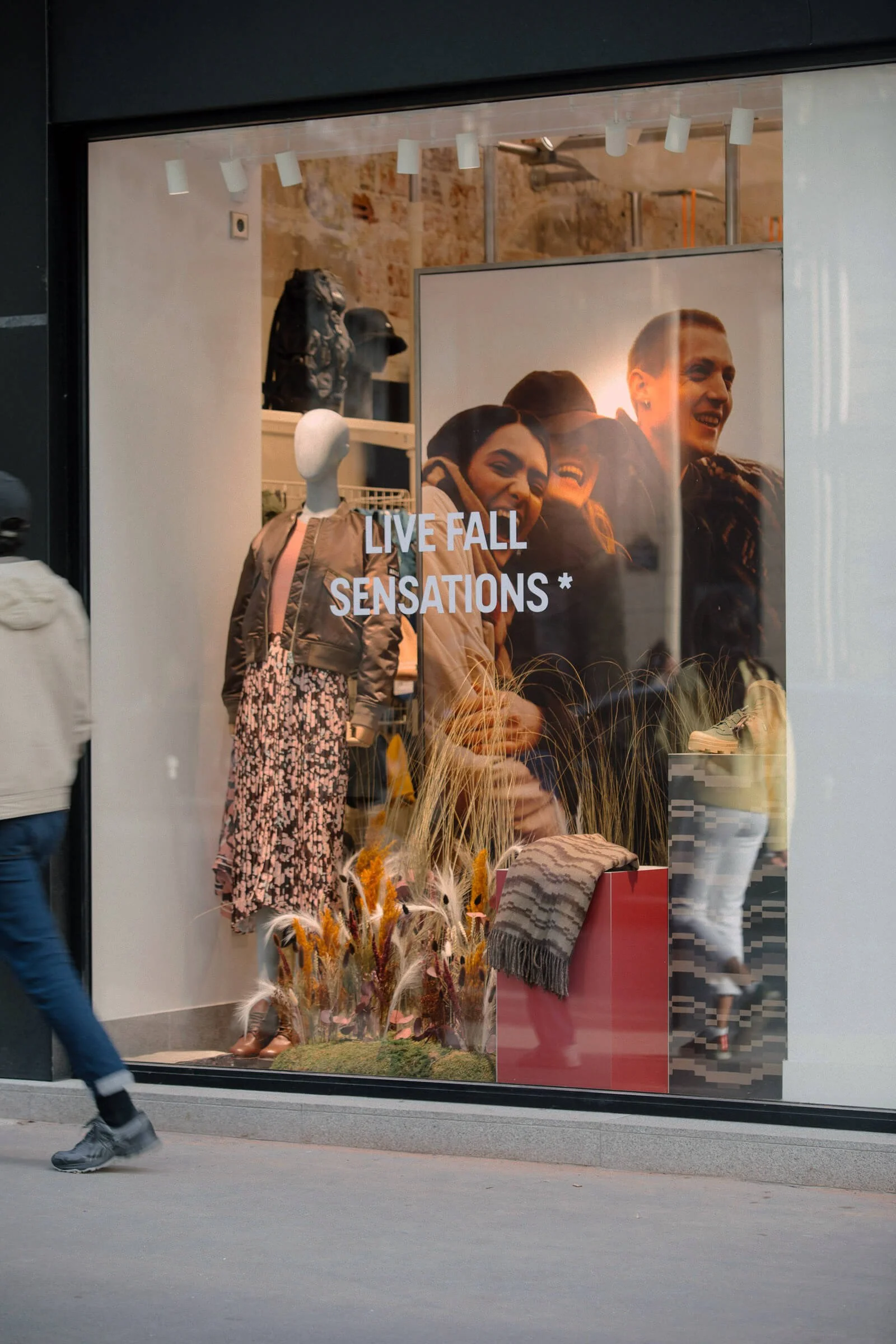 Vitrine de boutique avec mannequin habillé d'une jupette à fleur, accessoires, vêtements d'automne et un panneau annonçant « LIVE FALL SENSATIONS » avec une photo de trois personnes souriantes.