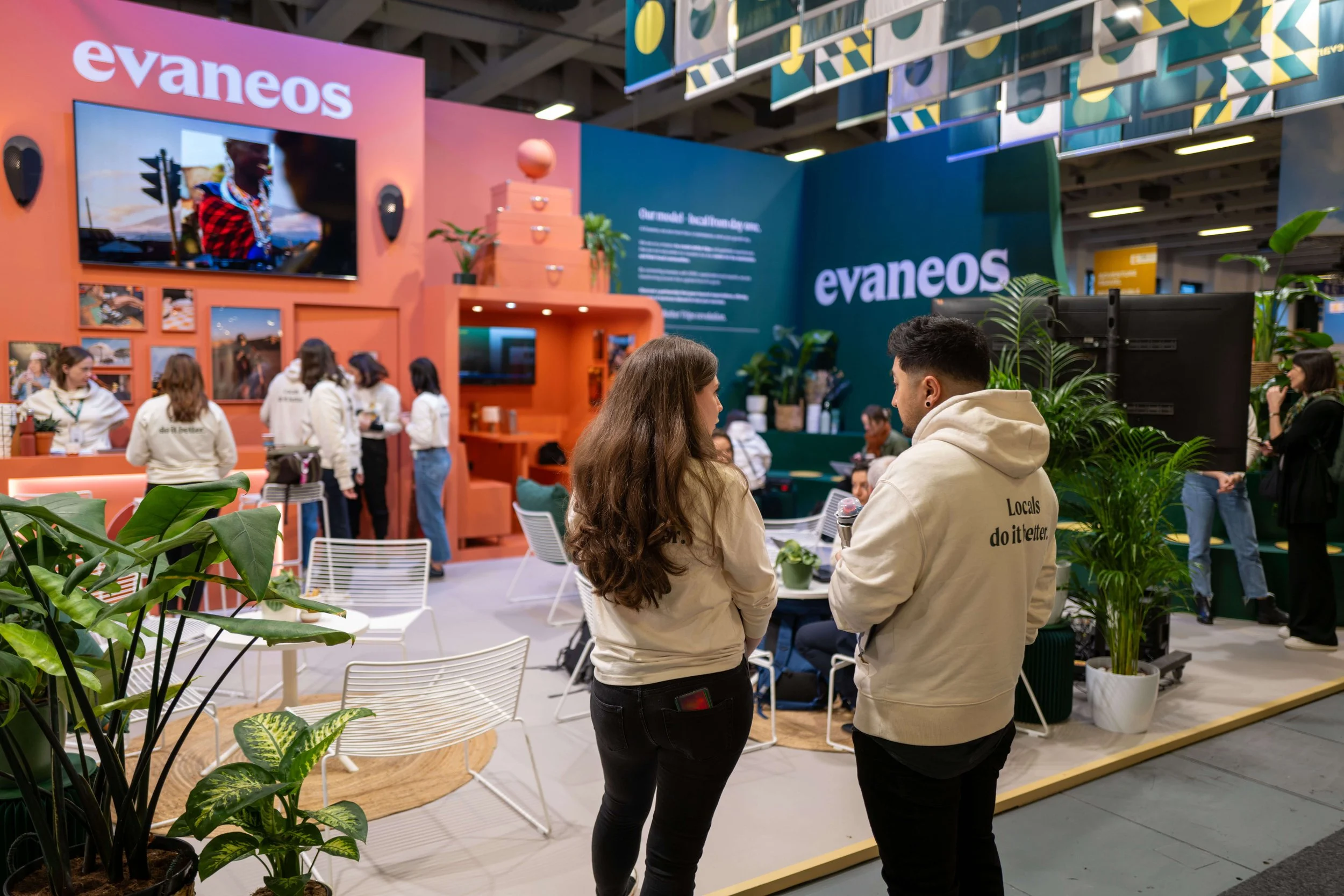 Deux personnes discutent dans un stand d'exposition avec le logo « evaneos » en arrière-plan, plusieurs autres personnes interagissent autour de stands colorés et de plantes décoratives.