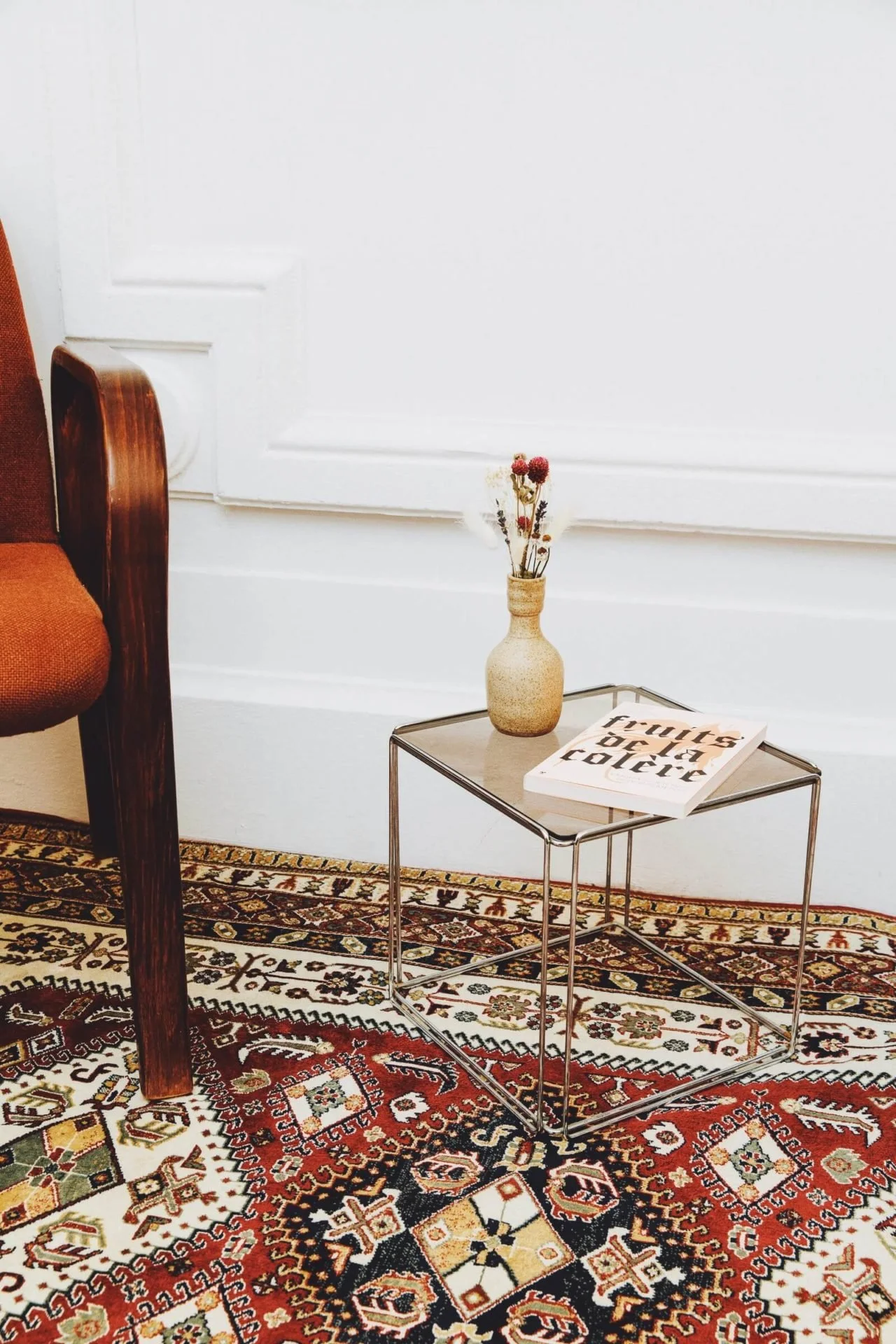 Coin de salon avec une chaise marron, une petite table en métal, un vase avec des fleurs séchées et un livre, posé sur un tapis à motifs colorés.