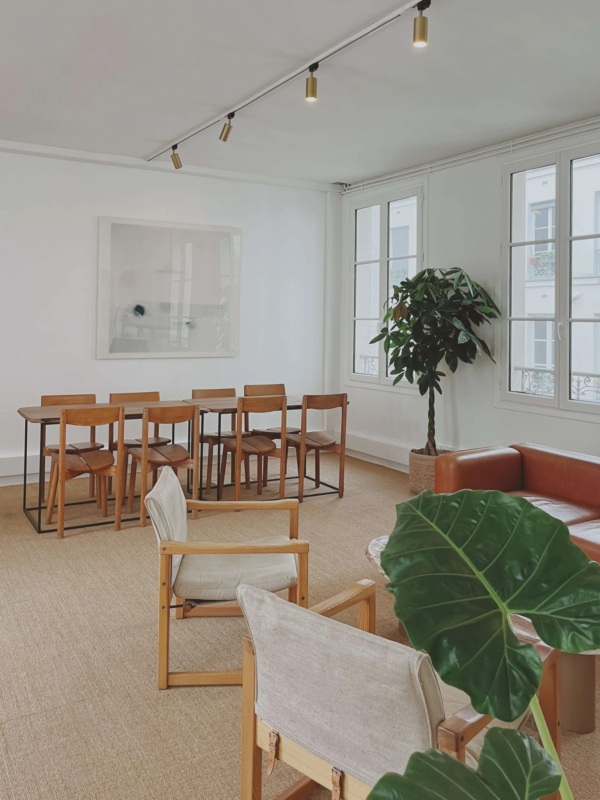 Salle de réunion moderne avec table en bois, chaises assorties, canapé en cuir, plantes vertes, fenêtres lumineuses et éclairage au plafond.