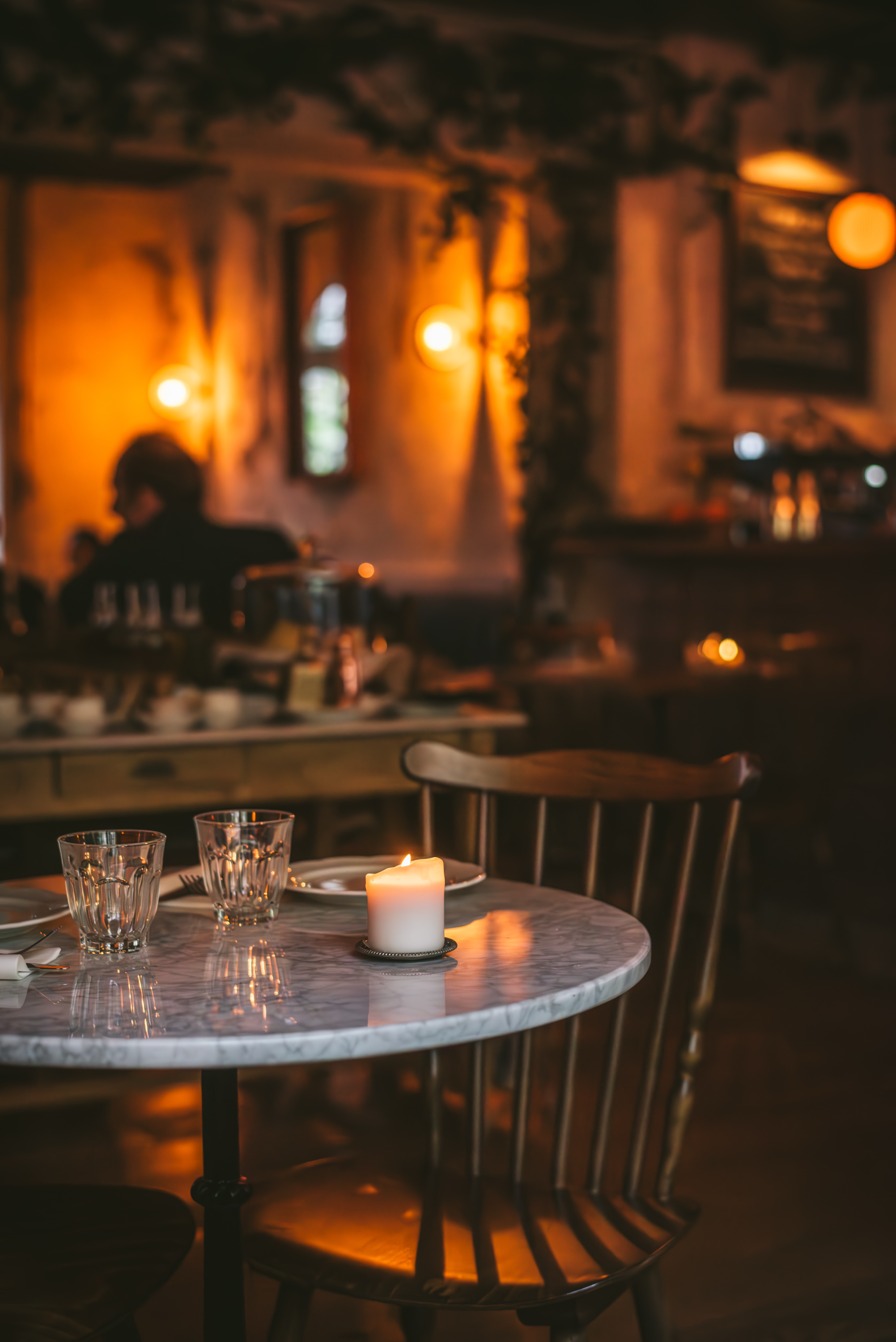 Table de restaurant avec une bougie allumée, 3 verres, une assiette, couverts et une nappe en marbre, ambiance chaleureuse avec lumière tamisée, dans un décor intérieur rustique.