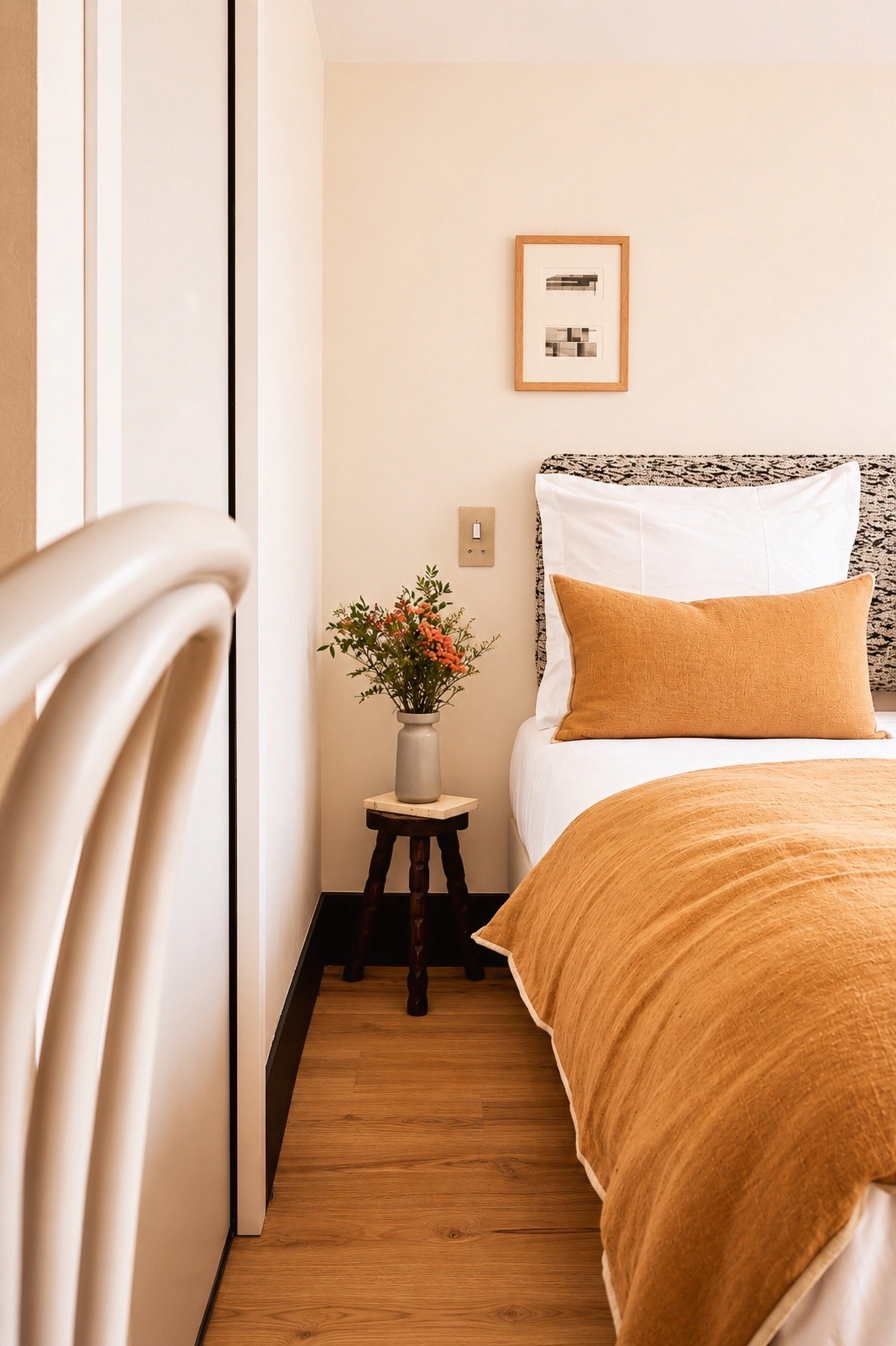 Une chambre avec un lit fait avec des draps de couleur beige, un coussin orange, une petite table en bois avec une vase contenant des fleurs, un tableau abstrait encadré en bois sur le mur, et un radiateur blanc au premier plan.