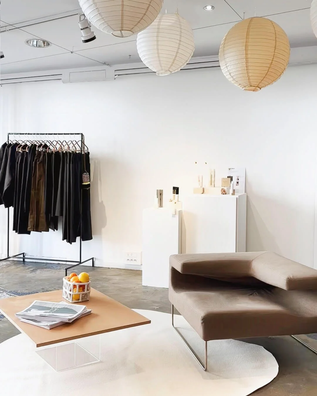 Intérieur d'une boutique ou d'un showroom avec un canapé beige, une table en bois avec des magazines et un panier de fruits, un portant avec des vêtements noirs, des lampions suspendus au plafond, et des présentoirs blancs avec des objets décoratifs.