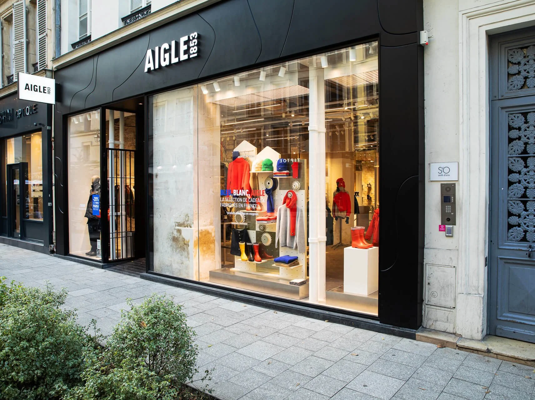 Vitrine d'un magasin de vêtements et accessoires avec des mannequins habillés en rouge, gris et vert, exposant des chaussures, des chapeaux et des vêtements, avec une façade noire et des fenêtres larges.