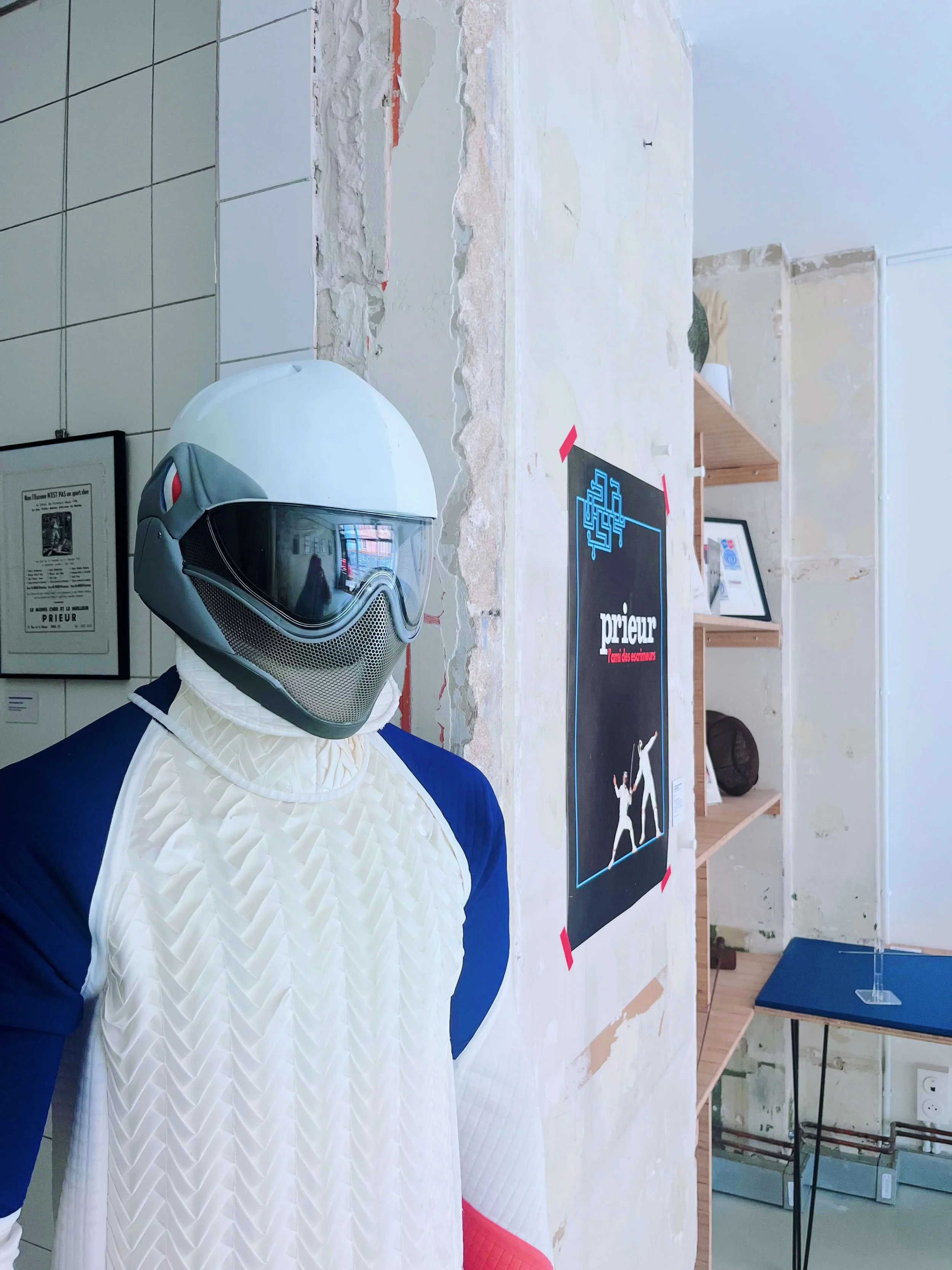 Une silhouette d'homme portant un casque de moto, un maillot de sport blanc avec des manches bleues, dans un espace intérieur avec des affiches et des étagères.