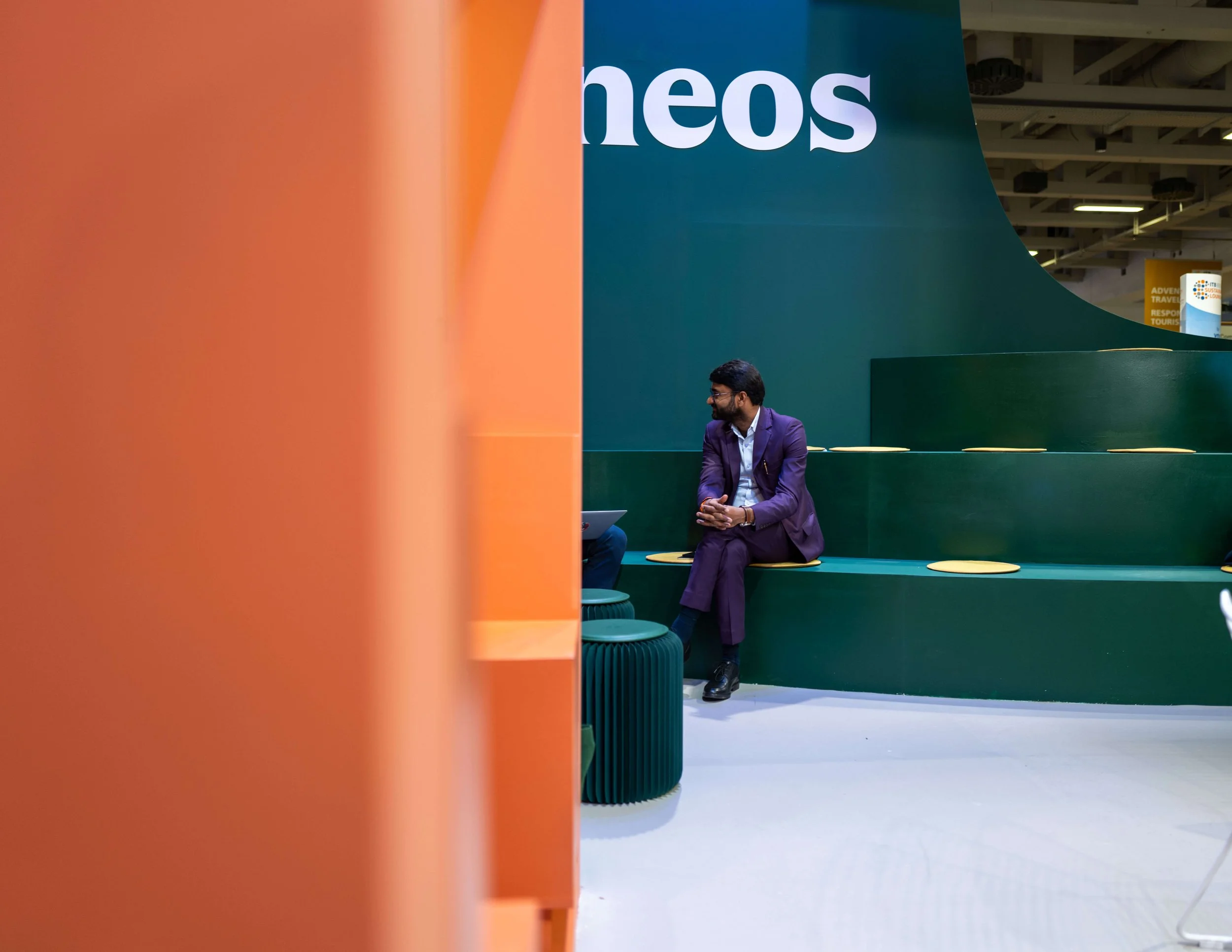 Un homme habillé en costume violet assis sur un banc vert dans une zone d'exposition avec un grand panneau en arrière-plan portant le mot 'theos'.