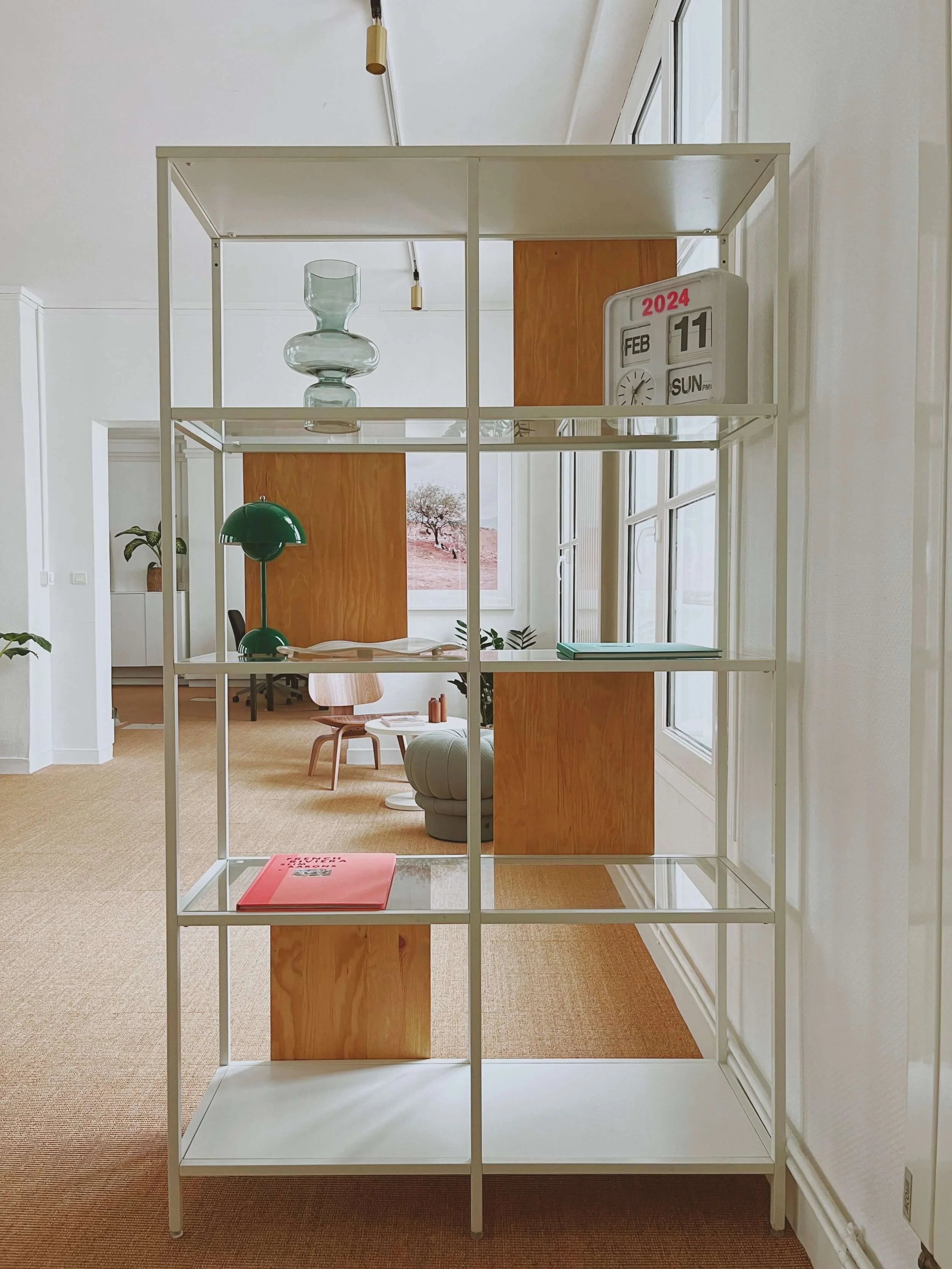 Salle moderne avec étagère blanche, vase en verre transparent, lampe verte, et calendrier indiquant le 11 février 2024. Mobilier et décoration minimalistes, ambiance chaleureuse et lumineuse.