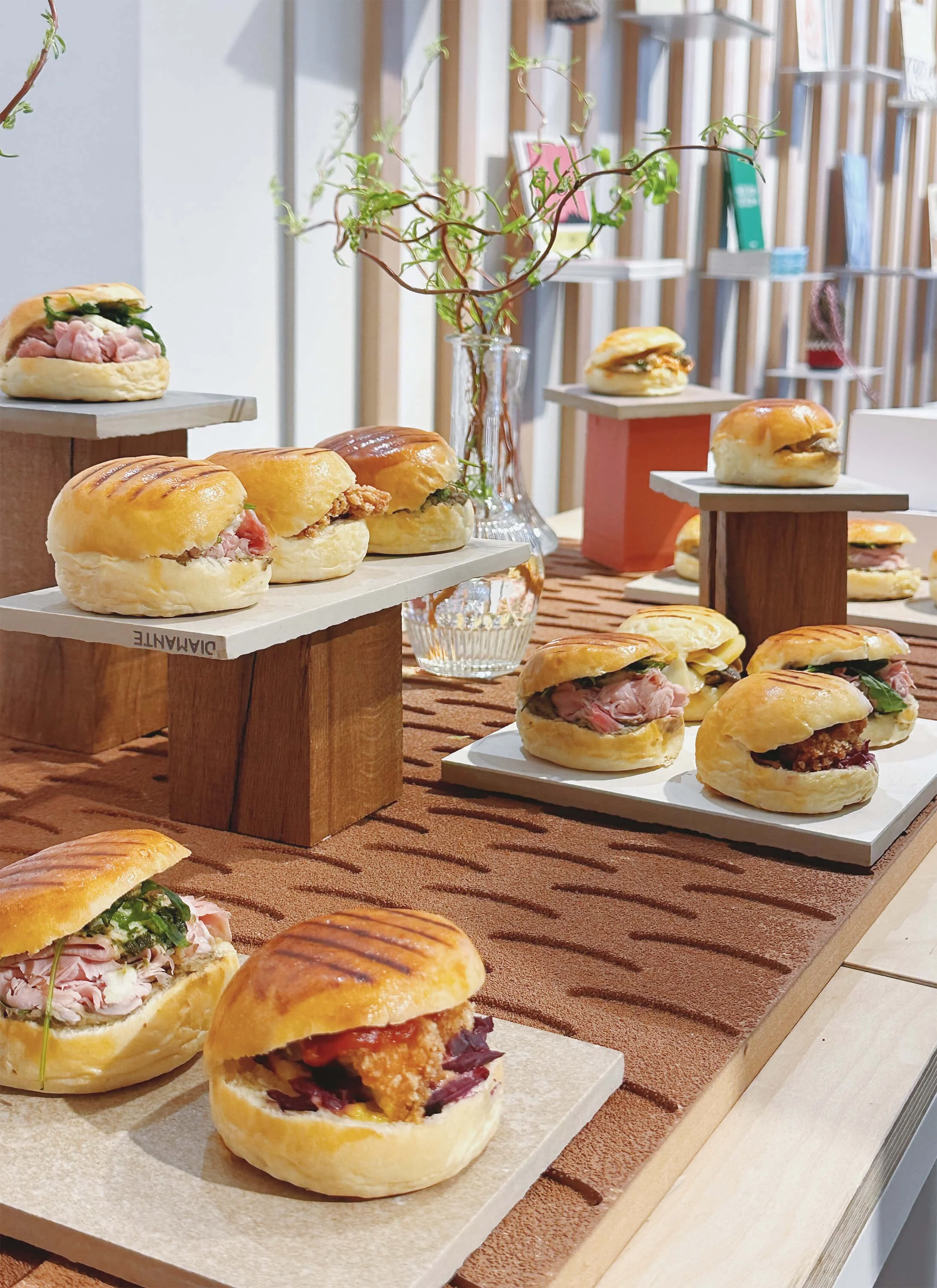 Une variété de petits sandwiches garnis de viandes, placés sur des plaques et des supports en bois sur une table avec un tapis marron et une vase avec une plante verte en arrière-plan.