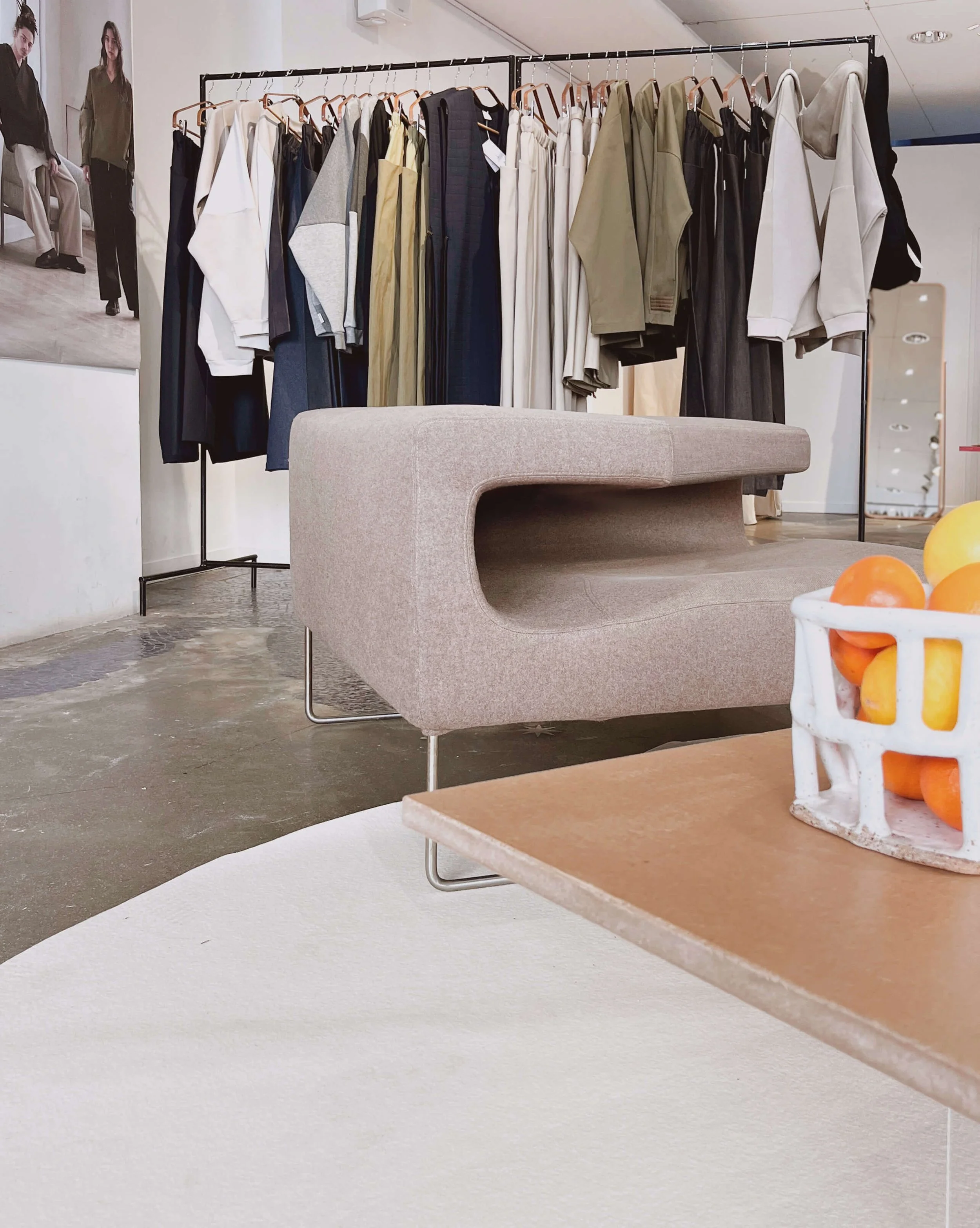 Vêtements suspendus sur une armoire dans une boutique ou un showroom, avec un banc beige et un panier de fruits oranges en premier plan.