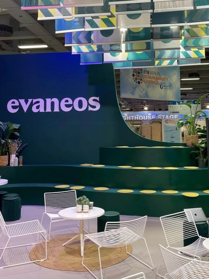 Espace talk du stand Evaneos pour le salon ITB Berlin dédié au Tourisme, avec des plantes, des fauteuils lounge blancs en métal et des tables basses qui on été loué, un mur avec le logo Evaneos stické en blanc, et des drapeaux suspendus au plafond.