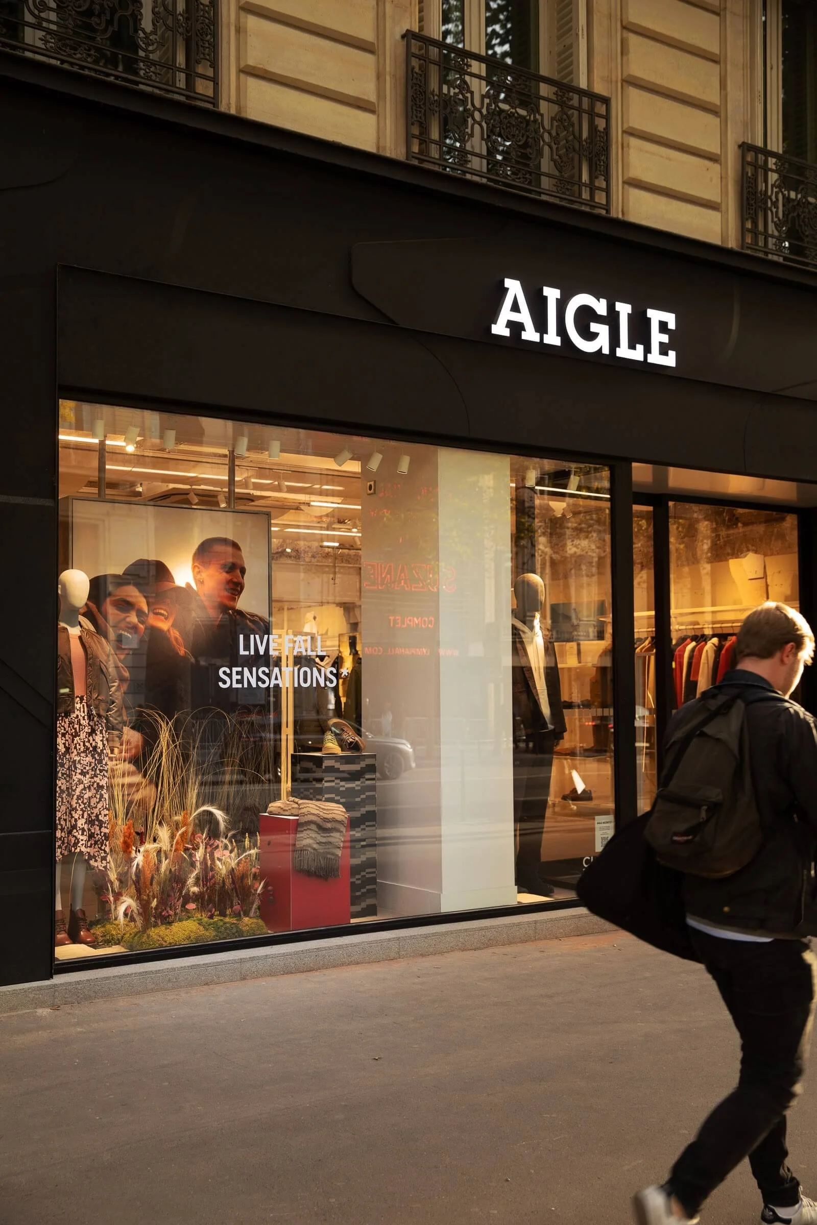Vitrine d'un magasin AIGLE avec mannequins, affichage de slogan "LIVE FALL SENSATIONS" et décor naturel avec des plantes et des vêtements en arrière-plan.