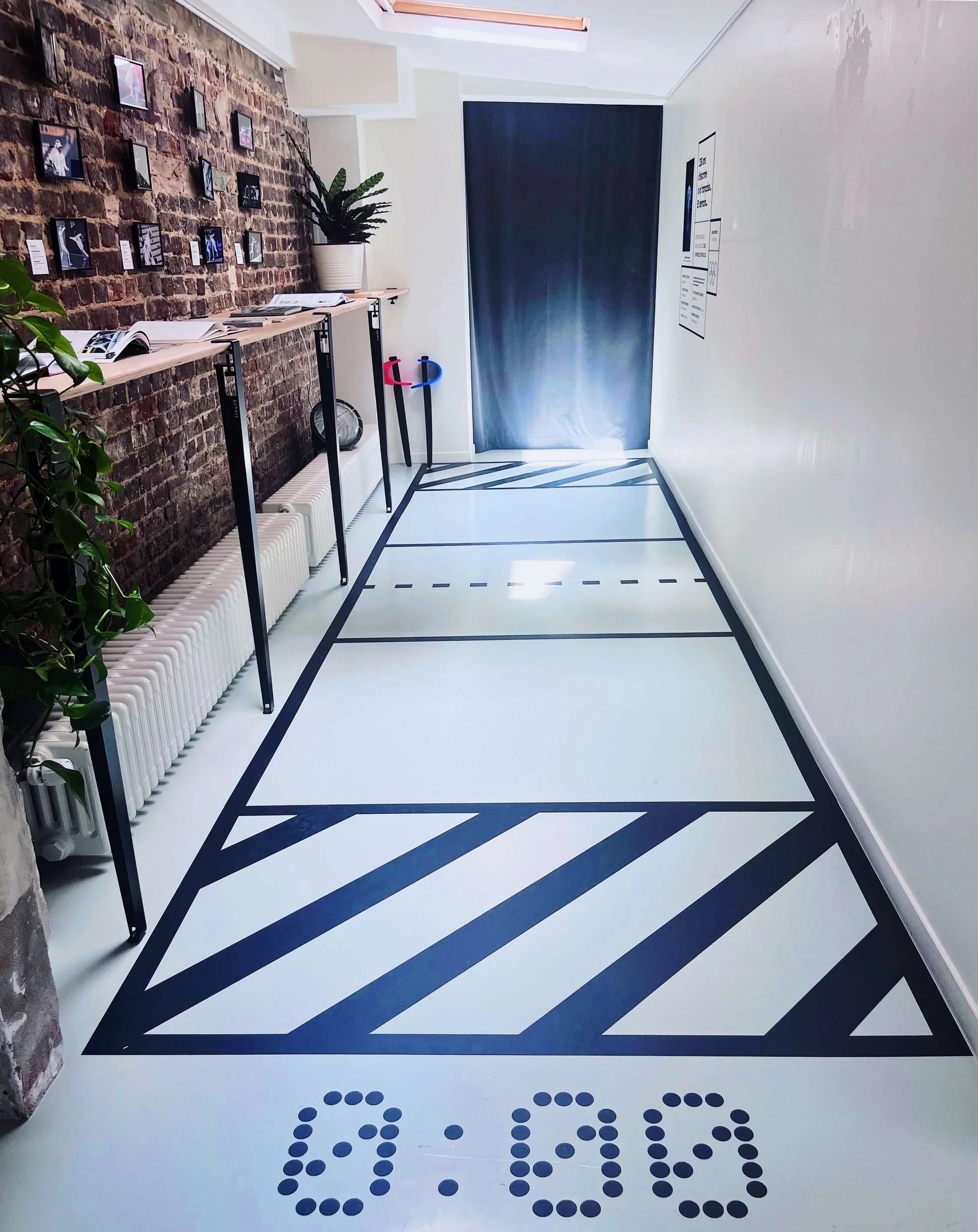 Espace de sport intérieur avec marquages au sol, tableau pour le basketball, plantes en pot et une façade en briques avec plusieurs photos encadrées.